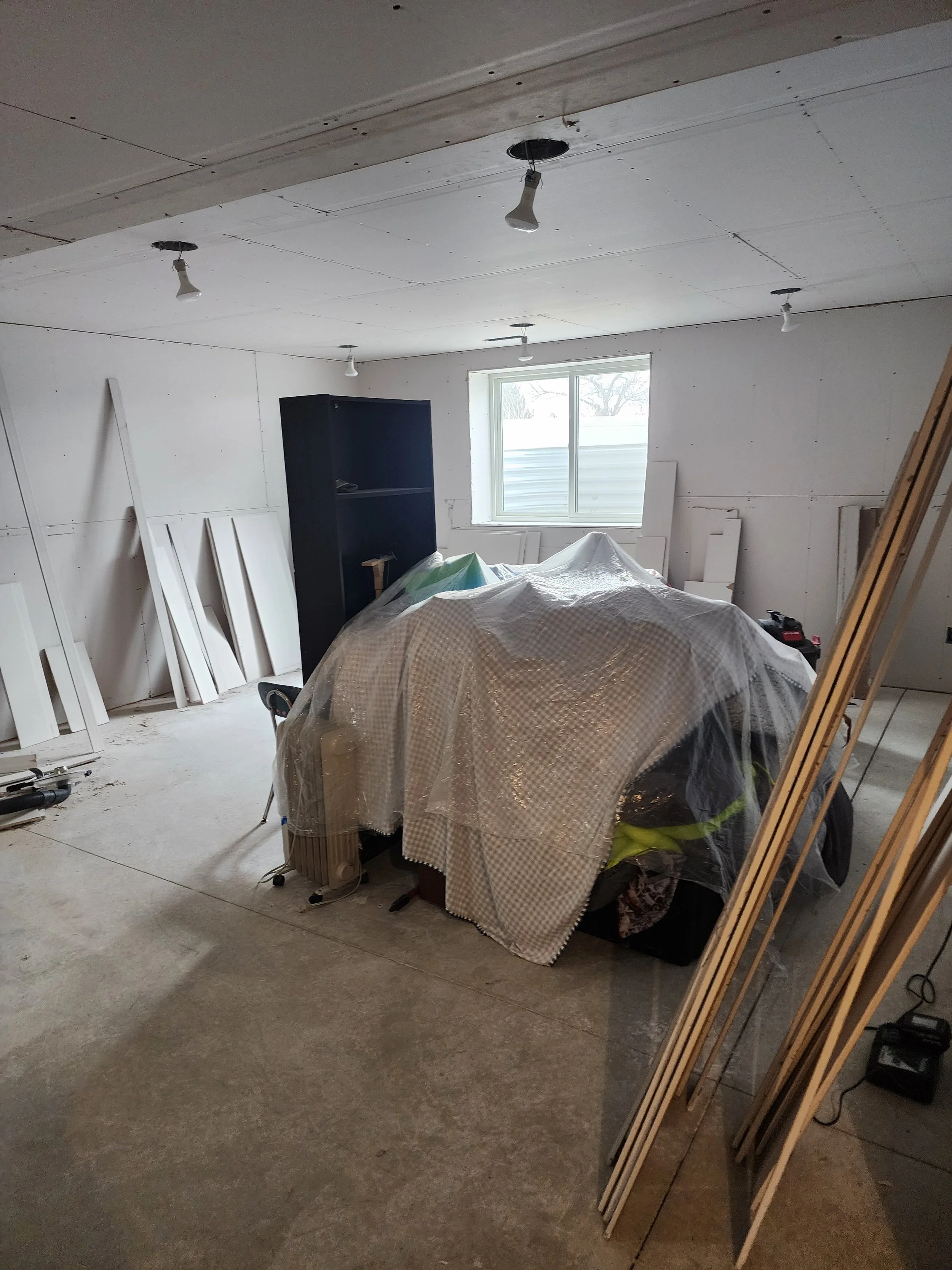 Room under construction with drywall on ceiling and walls, window, and construction materials covered with plastic sheeting.