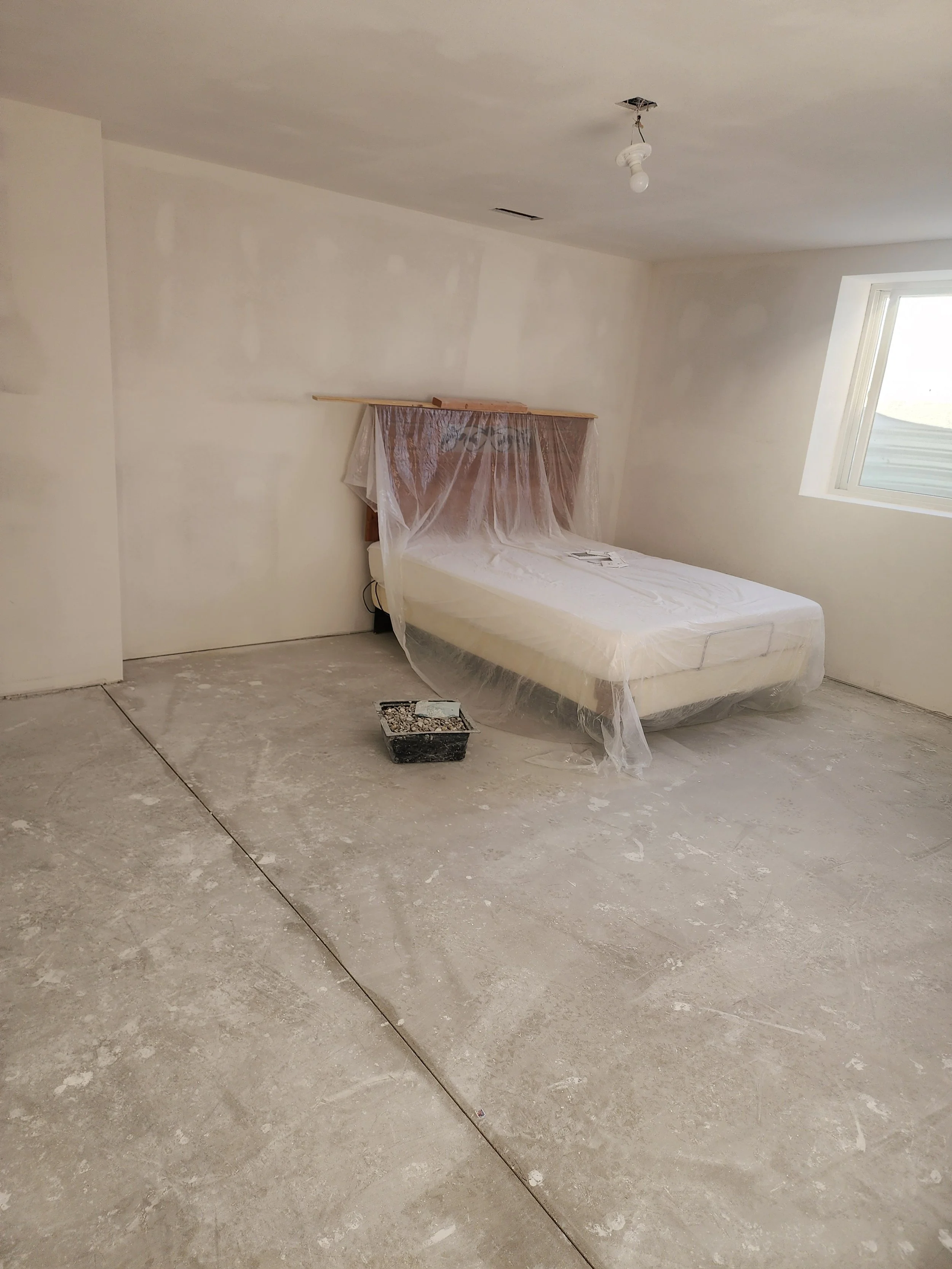 Bedroom under renovation with unfinished floor, a bed covered with plastic, a window, and a ceiling light fixture.