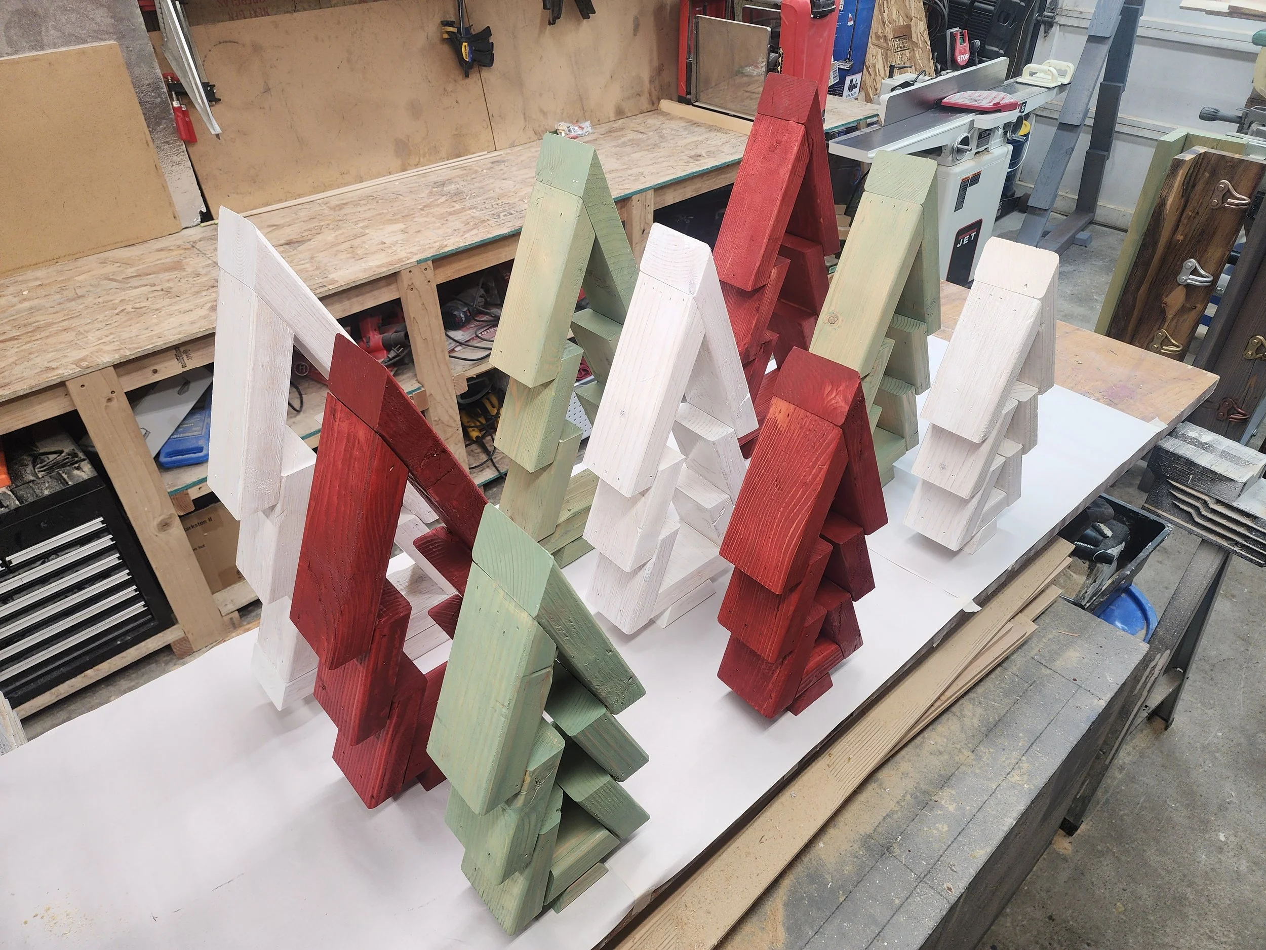 Colored wooden Christmas tree decorations in red, white, and light green, arranged on a workbench in a woodworking shop.