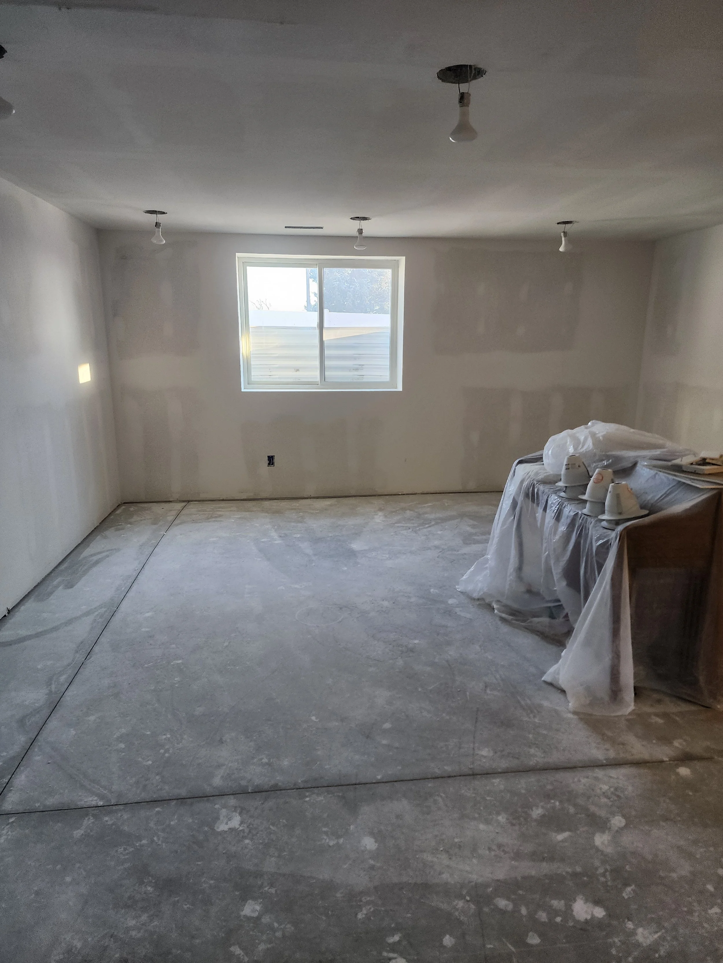 Unfinished room with drywall walls, exposed light bulbs, a window, and a table covered with plastic.