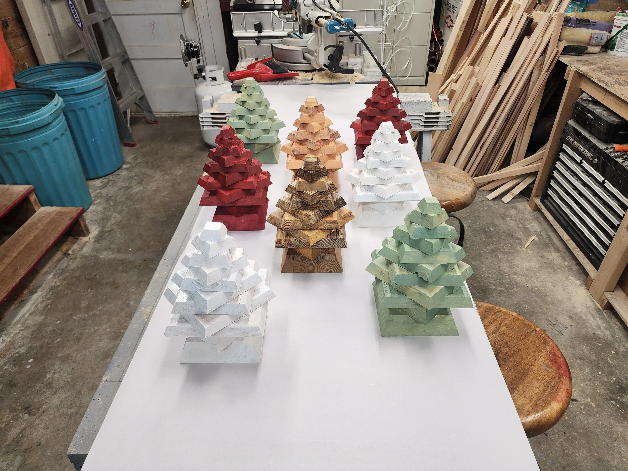 Colorful wooden Christmas tree ornaments arranged in rows on a white workbench in a woodworking workshop.