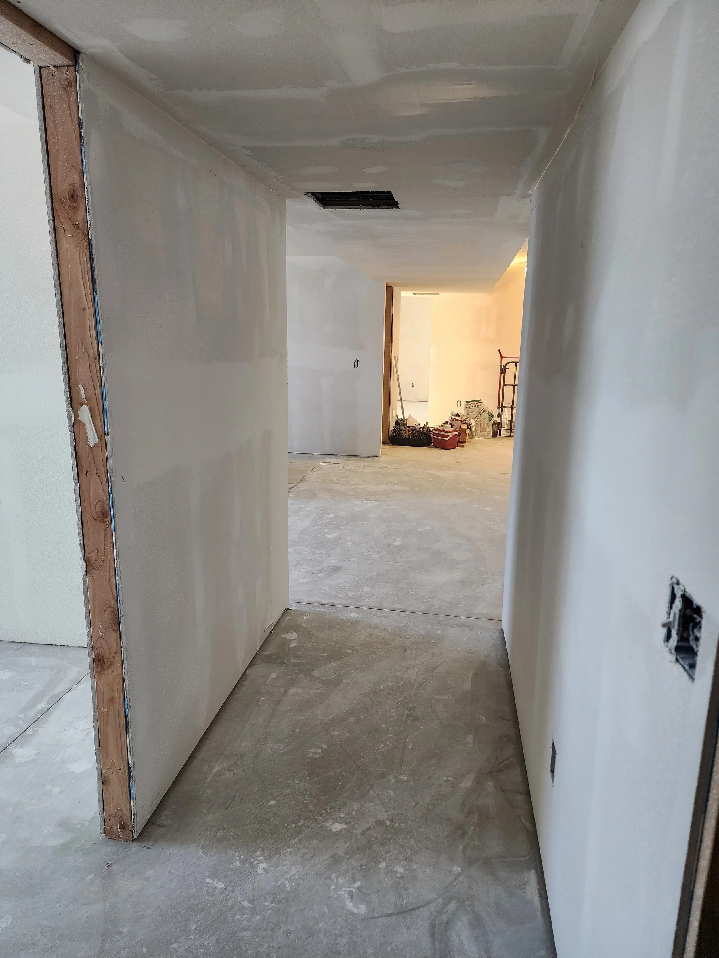 Interior view of a partially constructed or renovated house, showing finished drywall walls and ceiling, with construction tools and supplies visible in the background.