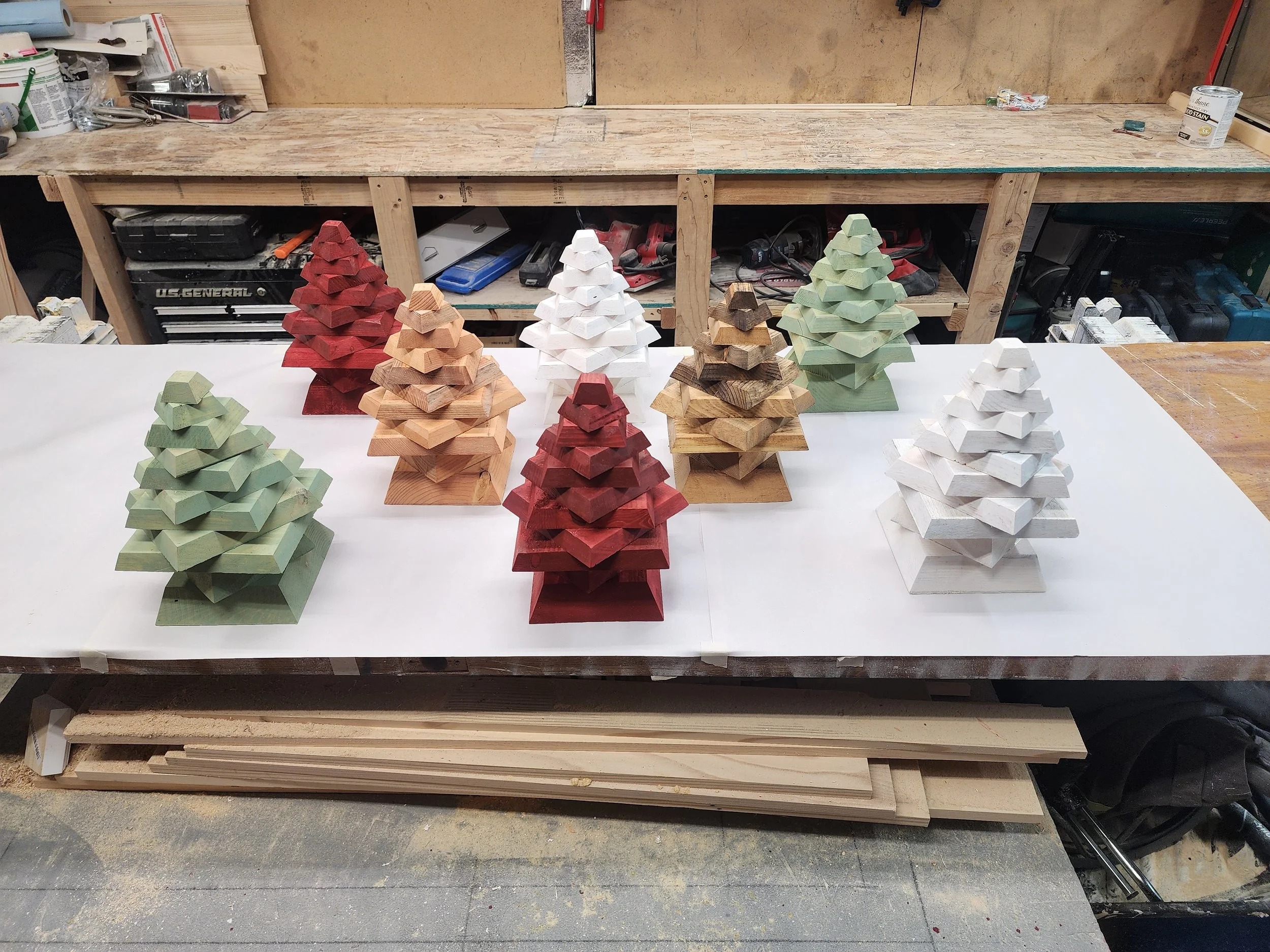 Several small wooden Christmas trees painted white, green, red, and natural wood color arranged on a workbench in a woodworking shop.