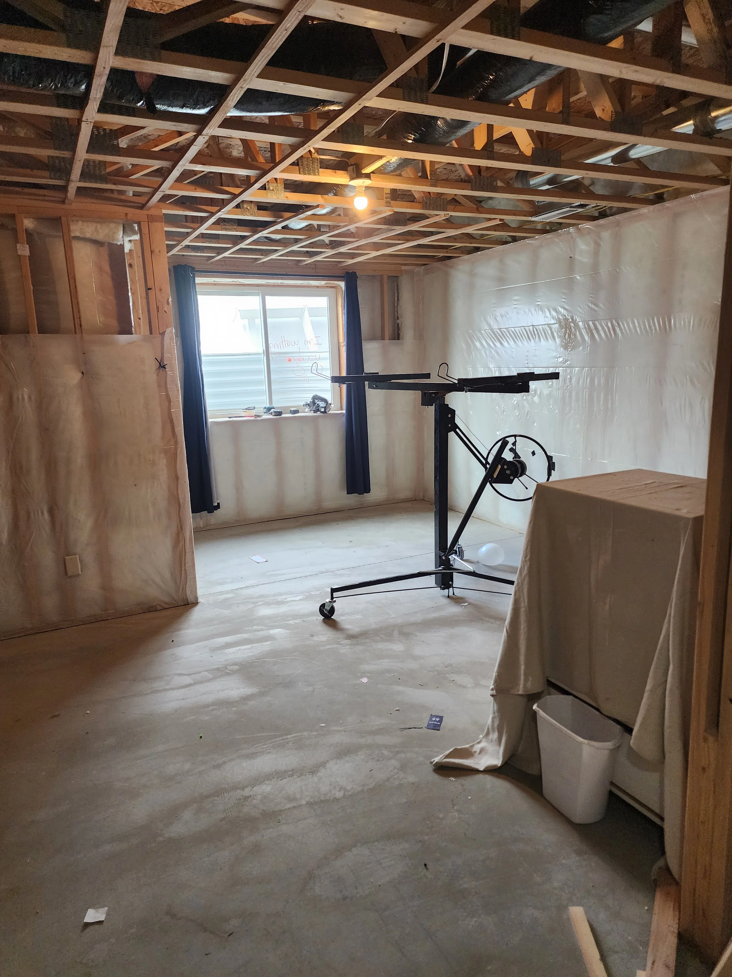 Room under construction with exposed wooden framing, a window with dark curtains, and a standing exercise bike in the center. There is construction material and a covered object on the right side.