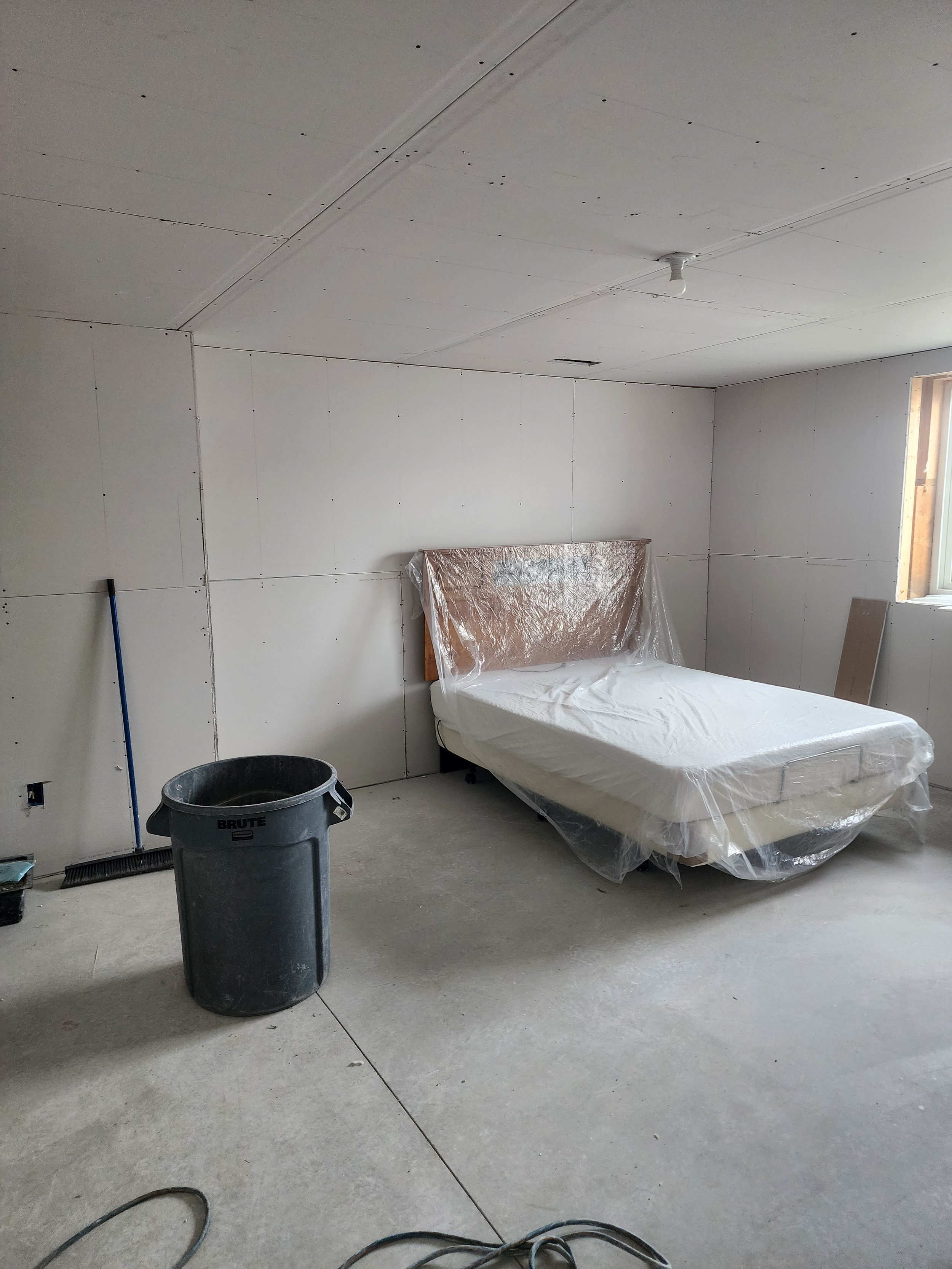 A bedroom under construction with bare drywall walls and ceiling, a mattress wrapped in plastic, a trash can, a broom, and a window letting in natural light.