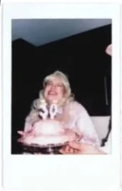 A person with long blonde hair wearing a light-colored top, smiling widely while celebrating a birthday with a pink frosted cake