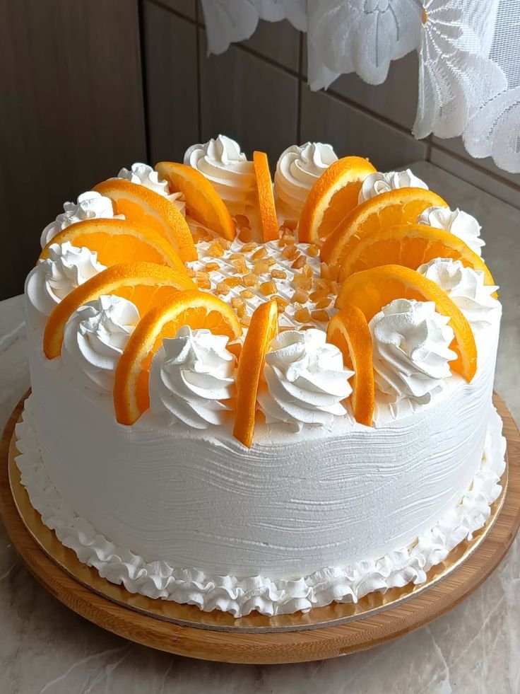 A round white cake decorated with whipped cream, orange slices, and small orange pieces, placed on a wooden cake board.