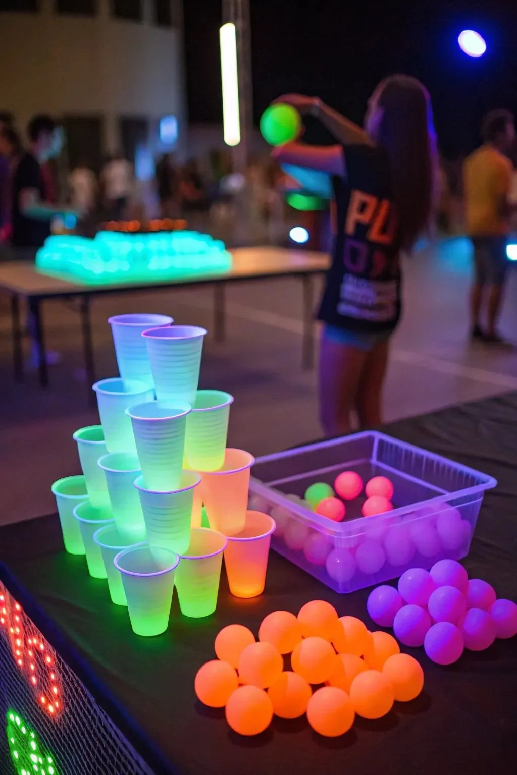 Glow-in-the-dark cups and balls at an arcade or carnival game booth at night, with people playing in the background.