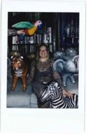 A woman sitting among colorful animal sculptures, including a zebra, a tiger, and an elephant, in an indoor setting with a bookshelf in the background.