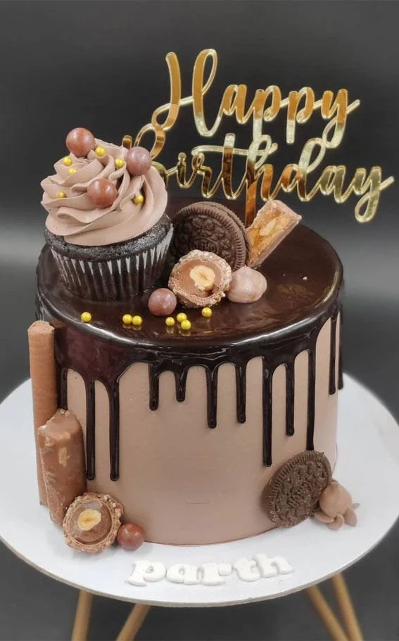Chocolate birthday cake with a "Happy Birthday" topper, decorated with chocolates, cookies, and gold and yellow ball sprinkles, on a white cake stand.