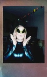 Person wearing an alien mask and shiny jacket, posing with hands up in a room with a string of colorful decorations.