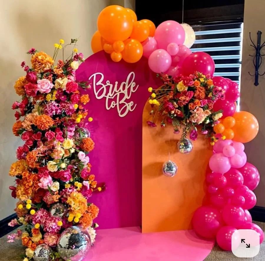 A decorative bridal shower backdrop featuring pink and orange balloons, colorful flower arrangements, and a pink sign that reads "Bride to Be."