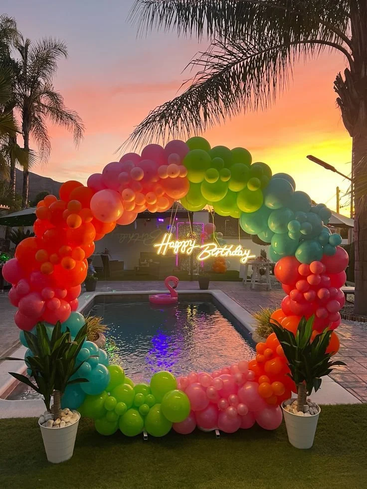 Colorful balloon arch with a 'Happy Birthday' neon sign over a swimming pool at sunset, decorated with potted plants and a pink flamingo float in the pool.