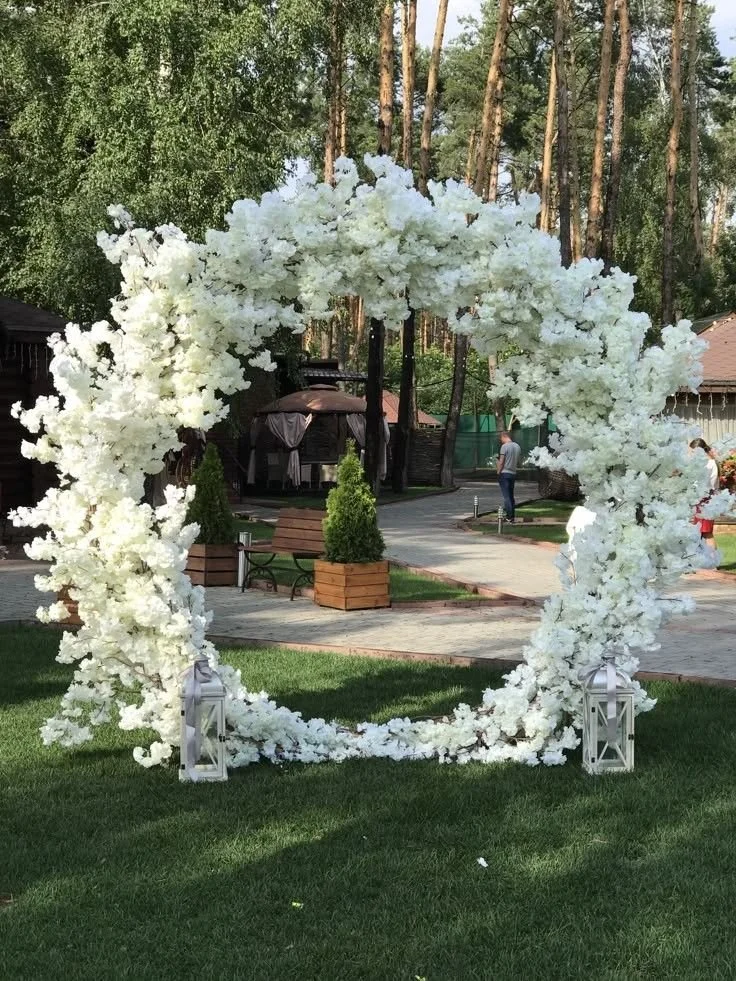 White floral arch decoration for outdoor event in a park or garden setting, with lanterns at the base.