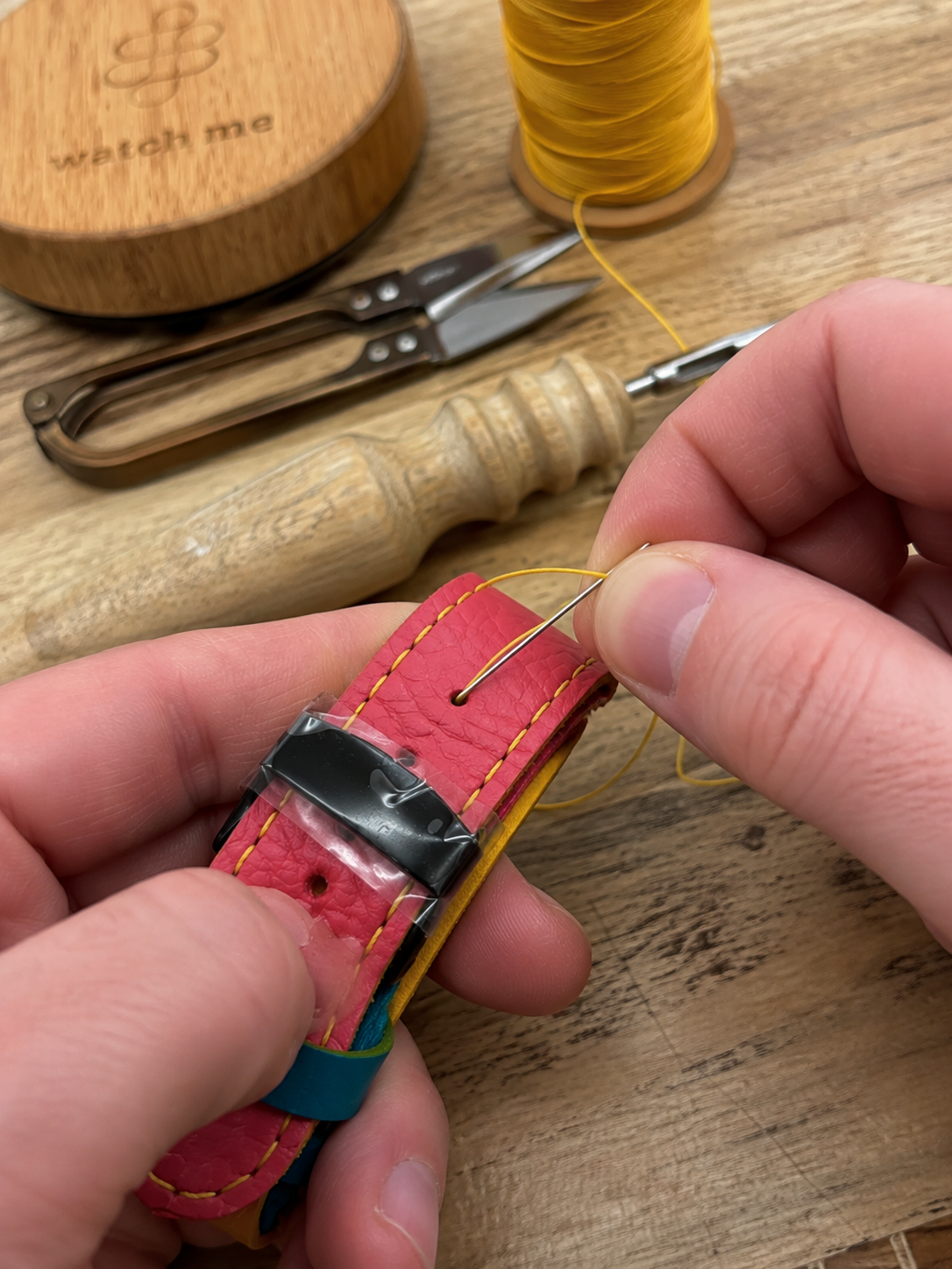 Une personne arrangeant une couture à la main sur une bande de cuir rose avec un fil jaune, avec des outils de couture en arrière-plan sur une surface en bois.