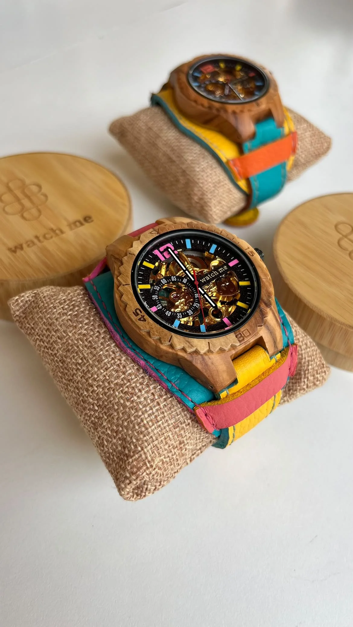 Deux montres colorées avec bracelets en tissu, exposées sur des coussins en toile de jute, avec deux boîtes en bois portant le mot 'Watch' gravé.