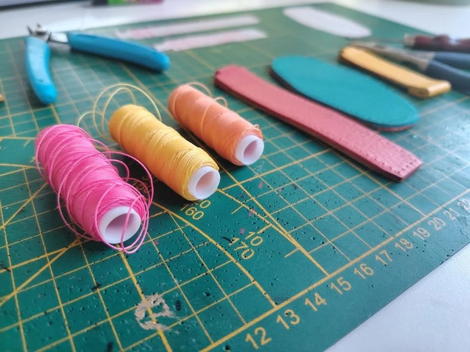 Matériel de couture sur une surface de travail : bobines de fil rose, jaune et orange, et diverses formes d'outils ou motifs en cuir ou en tissu dans différentes couleurs.
