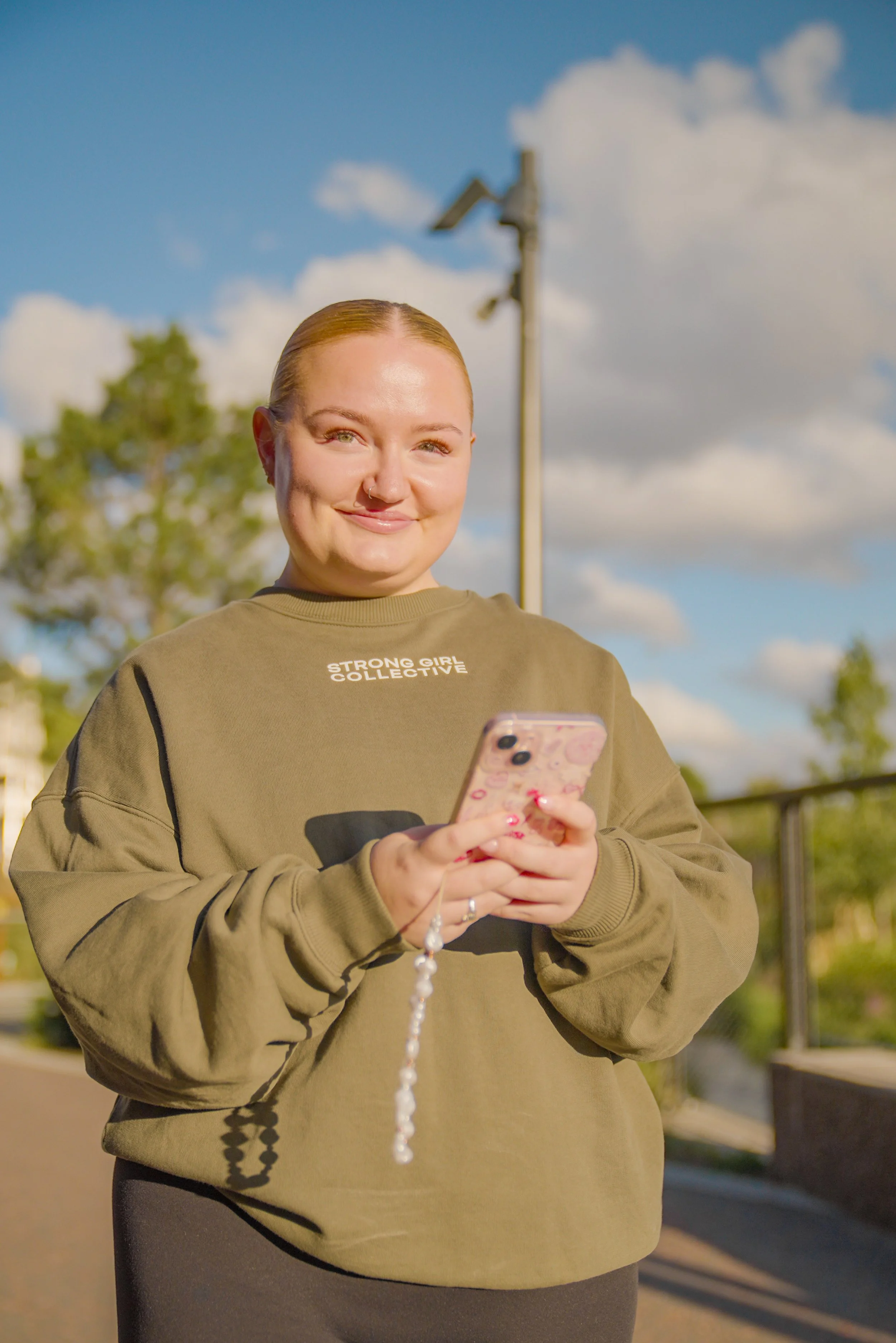 A young woman with blonde hair pulled back, smiling and looking at her phone, outdoors on a sunny day with clouds and trees in the background.