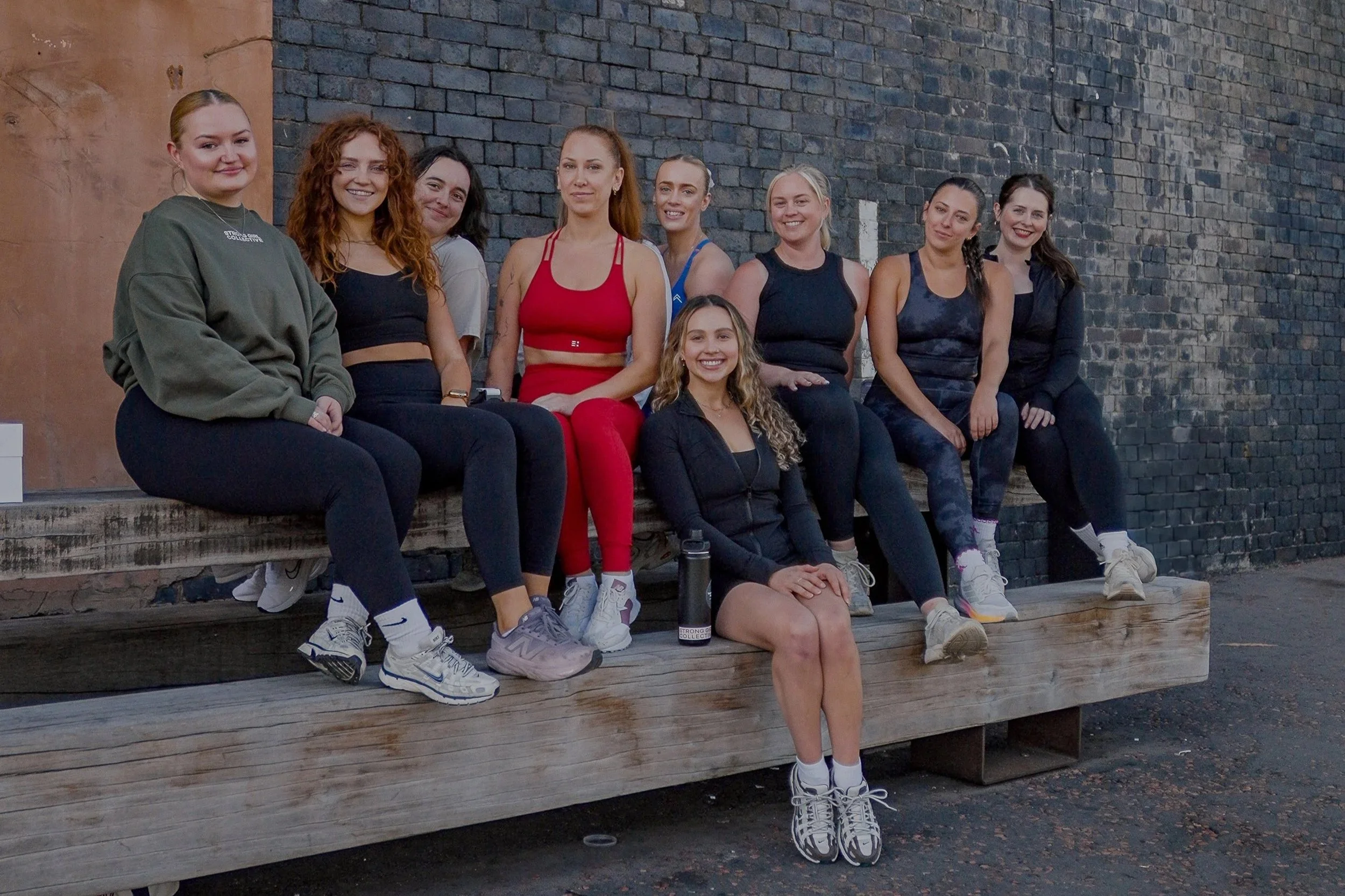 Group of ten women in athletic attire sitting on a wooden bench in front of a dark brick wall, smiling.