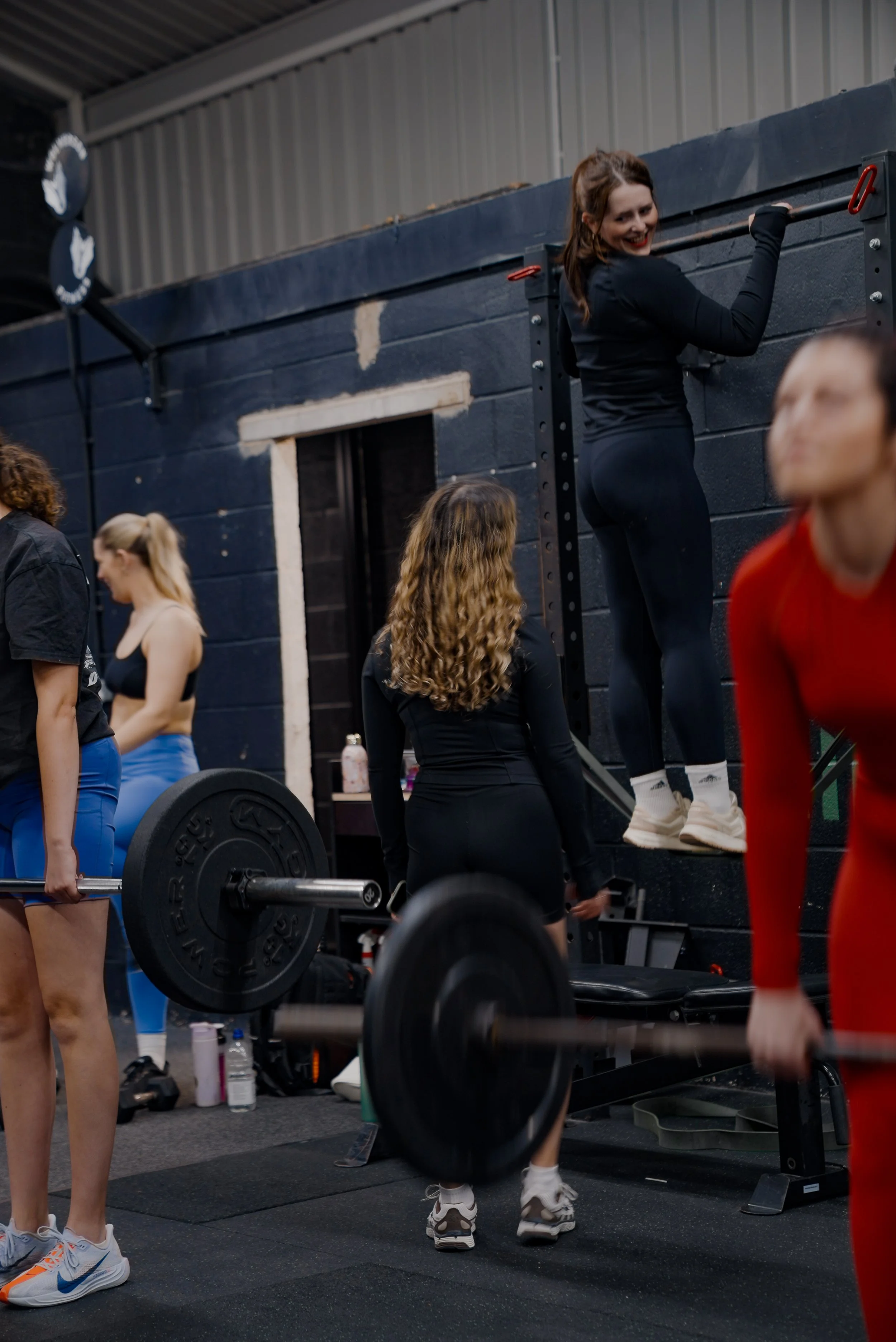 Women working out at a gym, some lifting weights, others doing pull-ups or hanging on a wall-mounted exercise bar, with gym equipment and a black wall in the background.