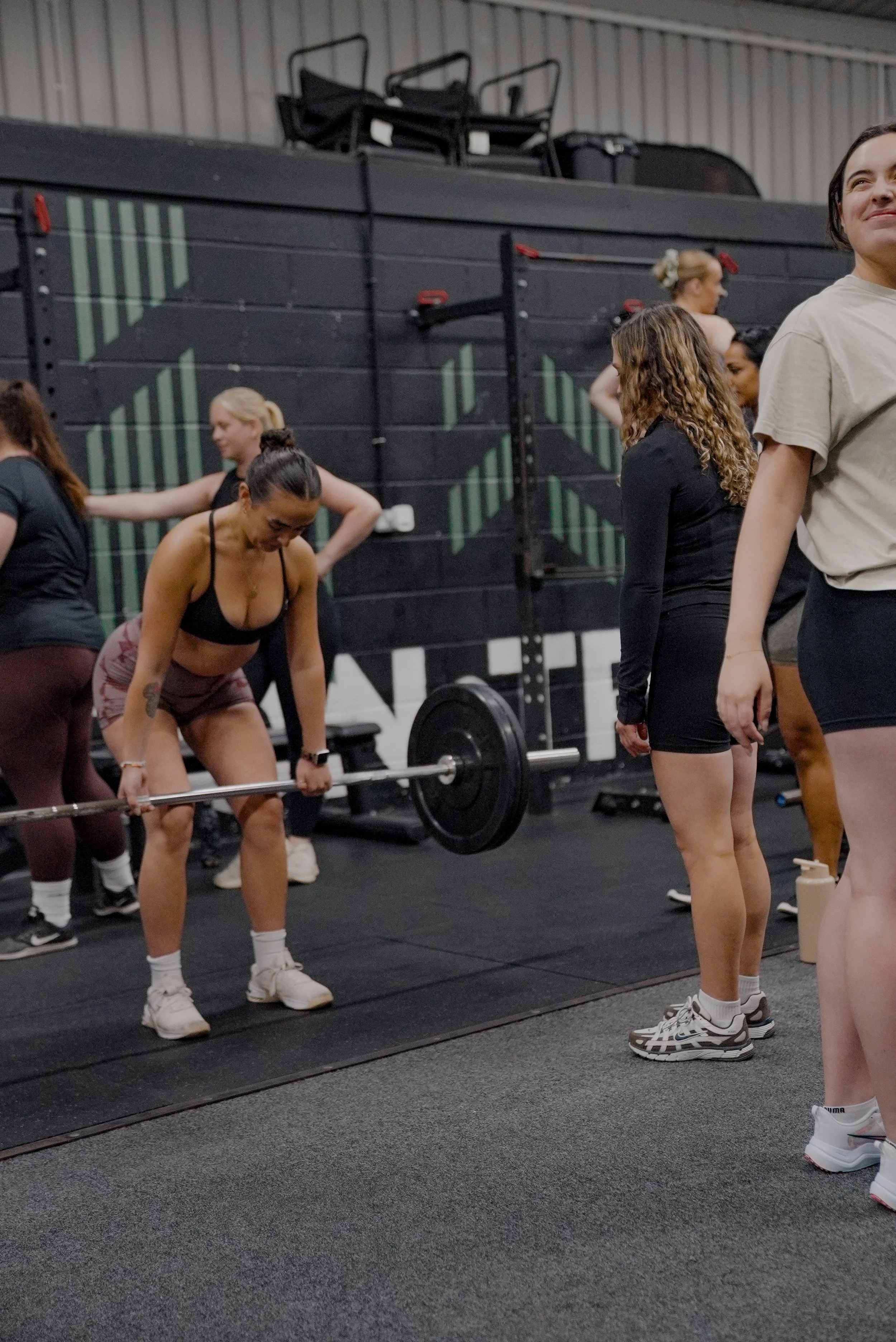 Women in a gym lifting weights and standing in a line, with a fitness wall and equipment in the background.