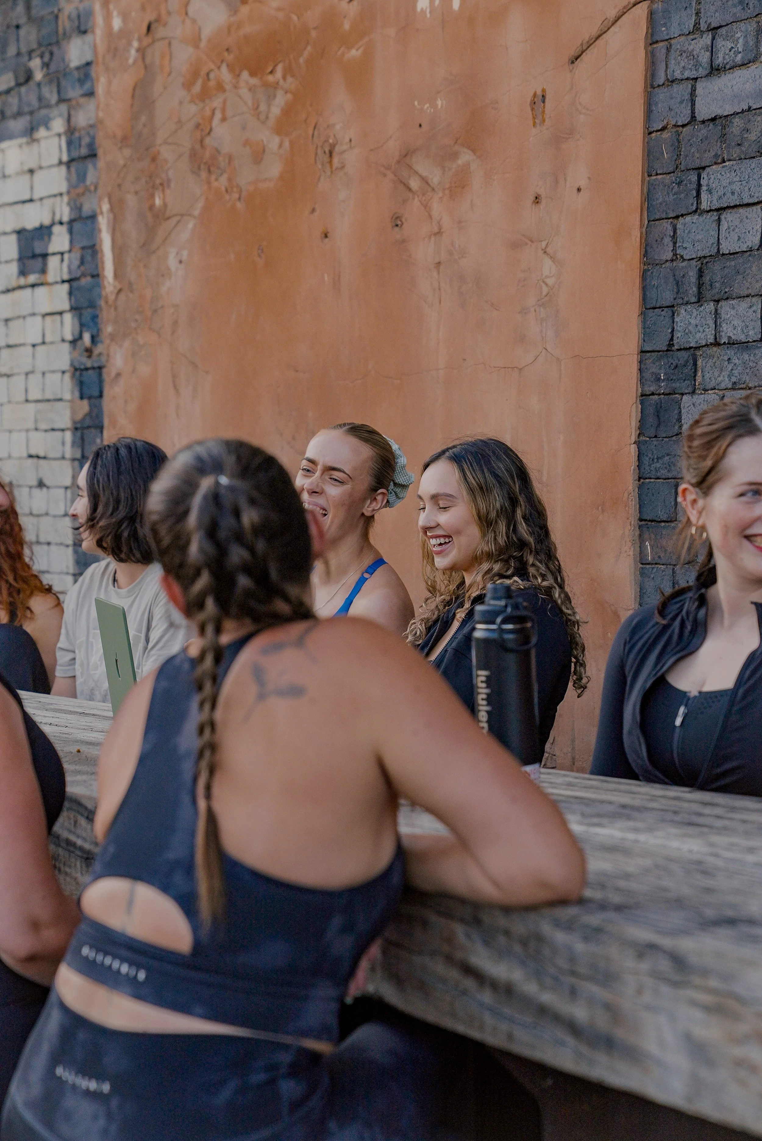 A group of women sitting at a wooden outdoor table, laughing and enjoying each other's company against an unfinished brick and concrete wall.