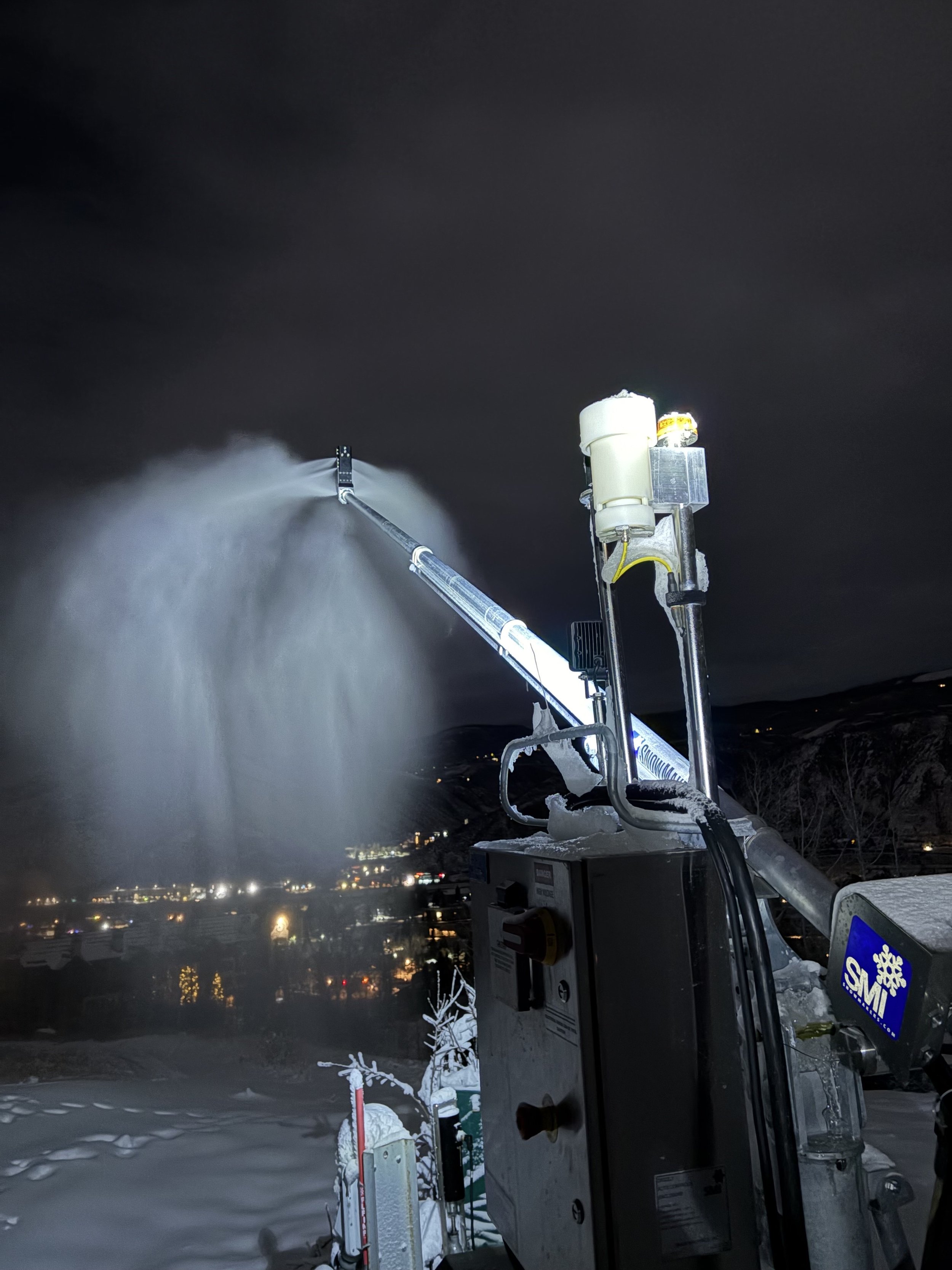 A snowmaking machine spraying artificial snow into the night sky, with snow-covered ground and distant city lights in the background.