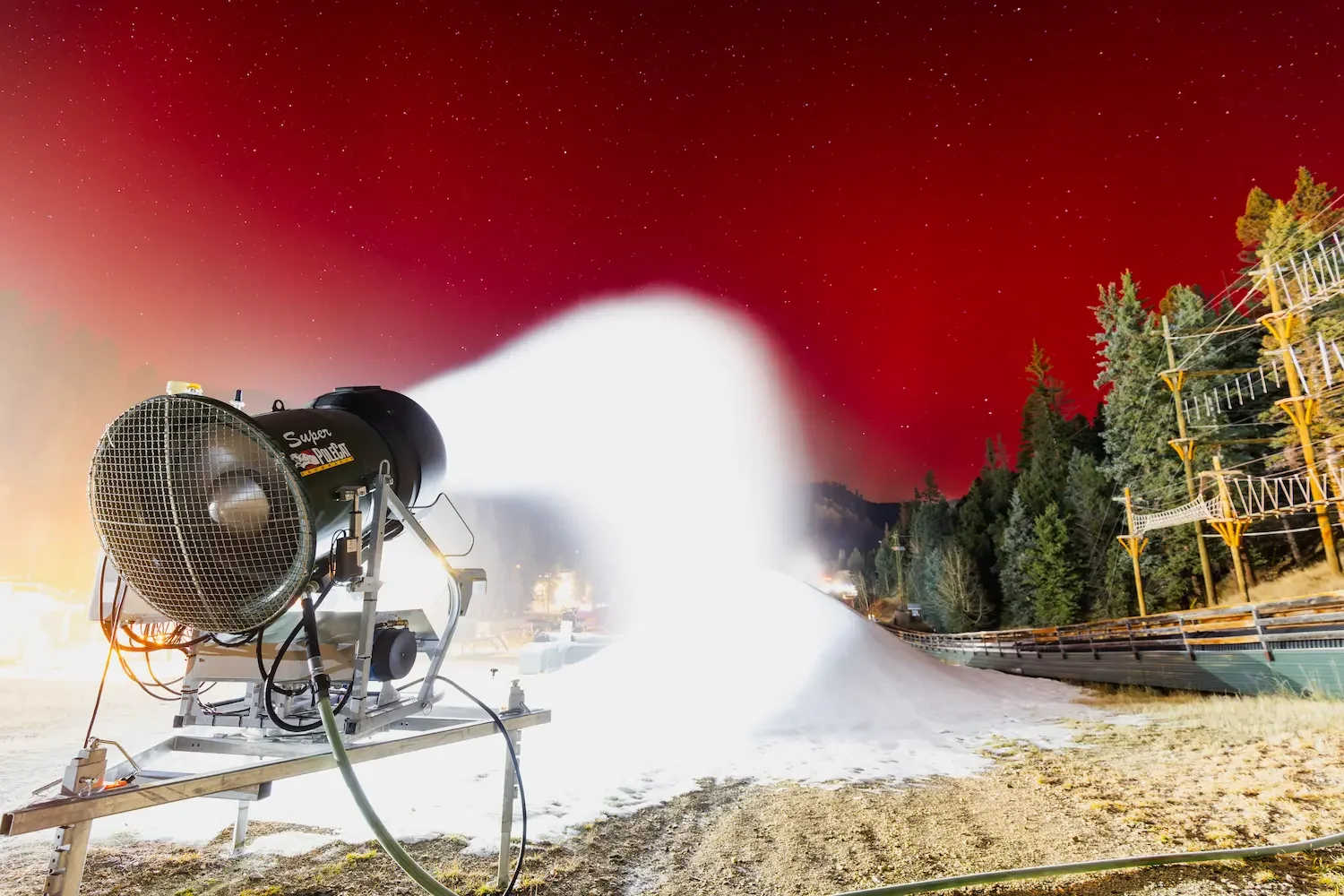 A snowmaking machine spraying artificial snow on a ski slope at night, with the sky glowing red and stars visible, and trees lining the side of the slope.