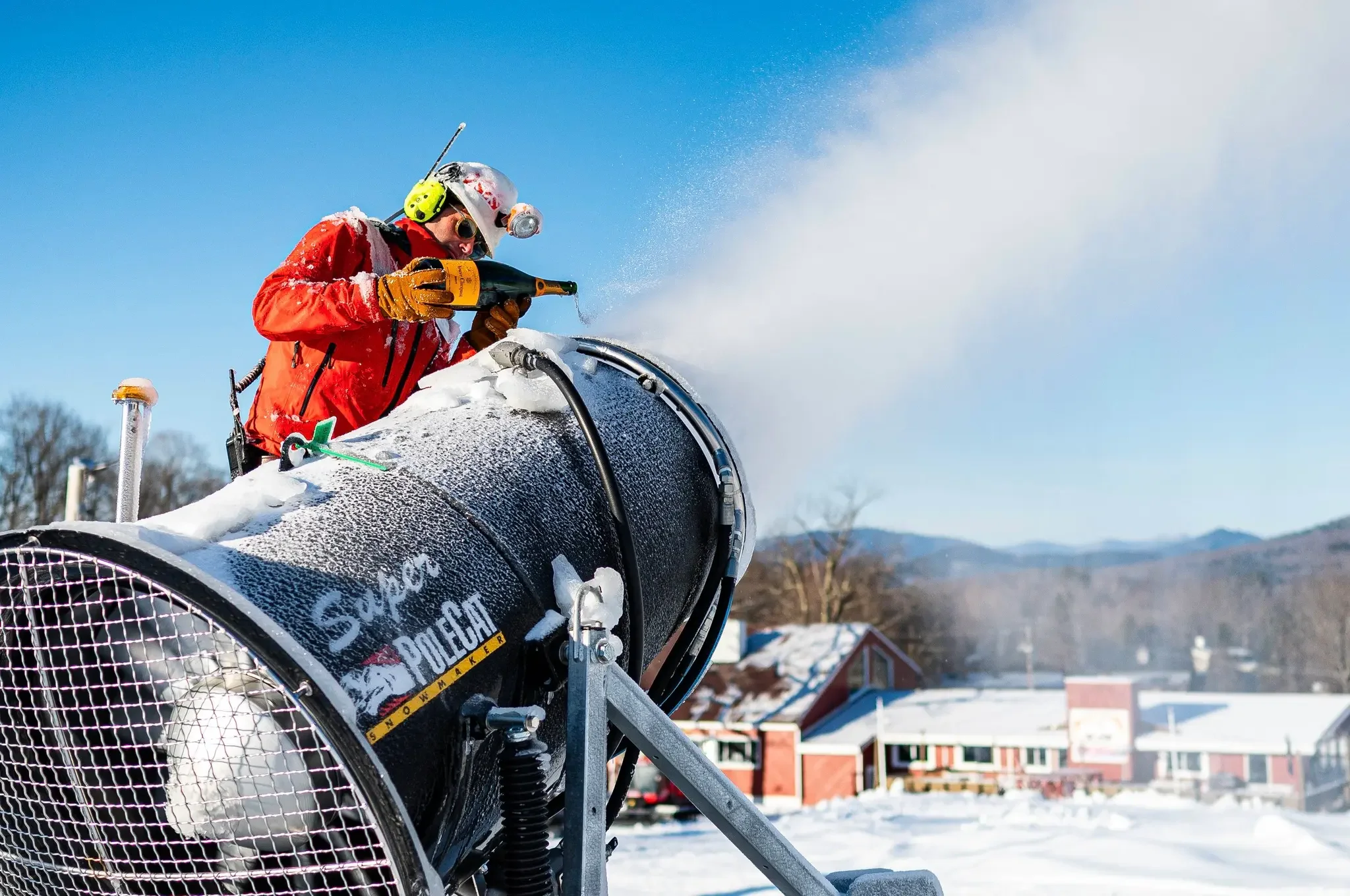 A man in a red jacket and safety equipment is spraying snow off a large snow-making machine outdoors on a snowy landscape with houses and mountains in the background.