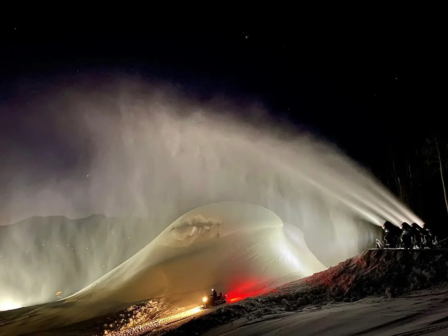 Nighttime scene of snow or ice being blown by a snowblower or snow plow device, with bright lights illuminating the snow and large snow drift, and a group of snow removal equipment on the right.