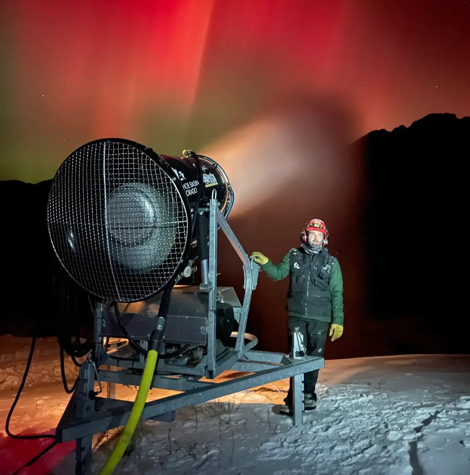 A person in winter gear, including a helmet and gloves, standing next to a large snowmaking machine on snow, with the Northern Lights visible in the night sky in the background.
