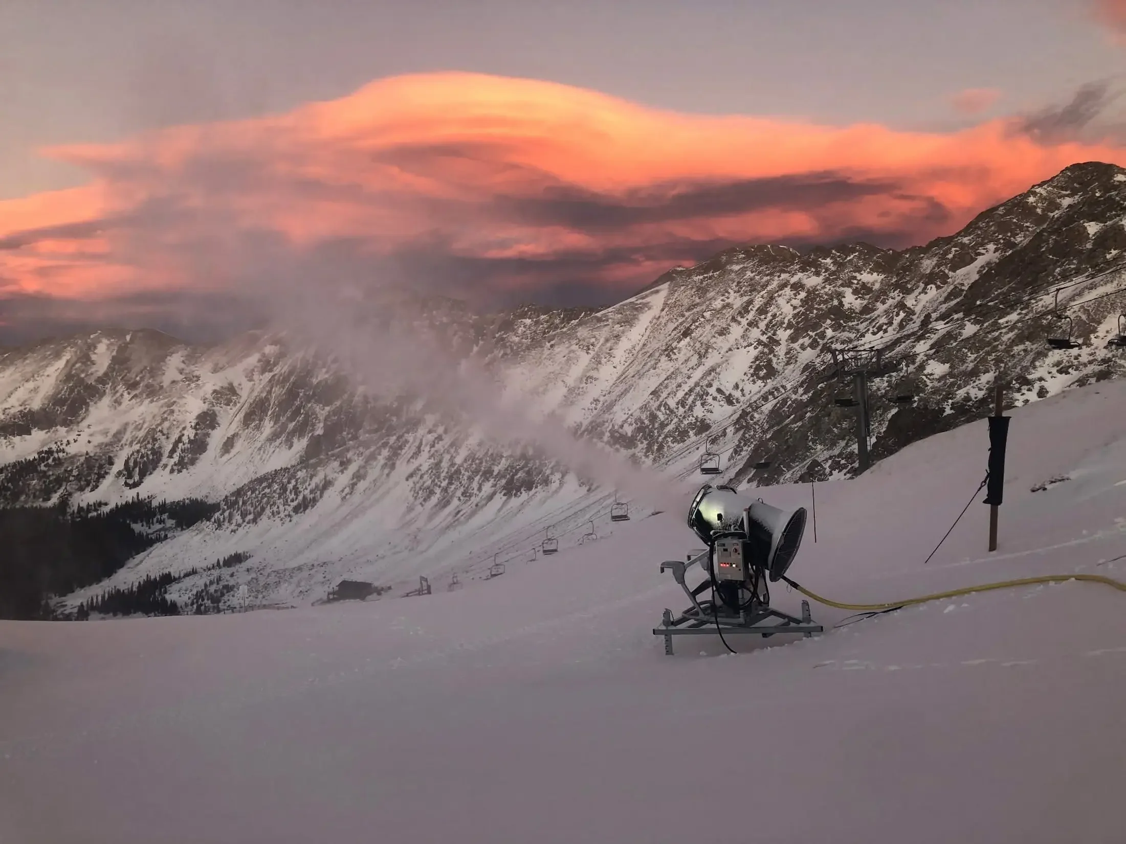 Snow-covered mountain landscape at sunset with ski resort equipment and chairlifts, pink and orange clouds in the sky.