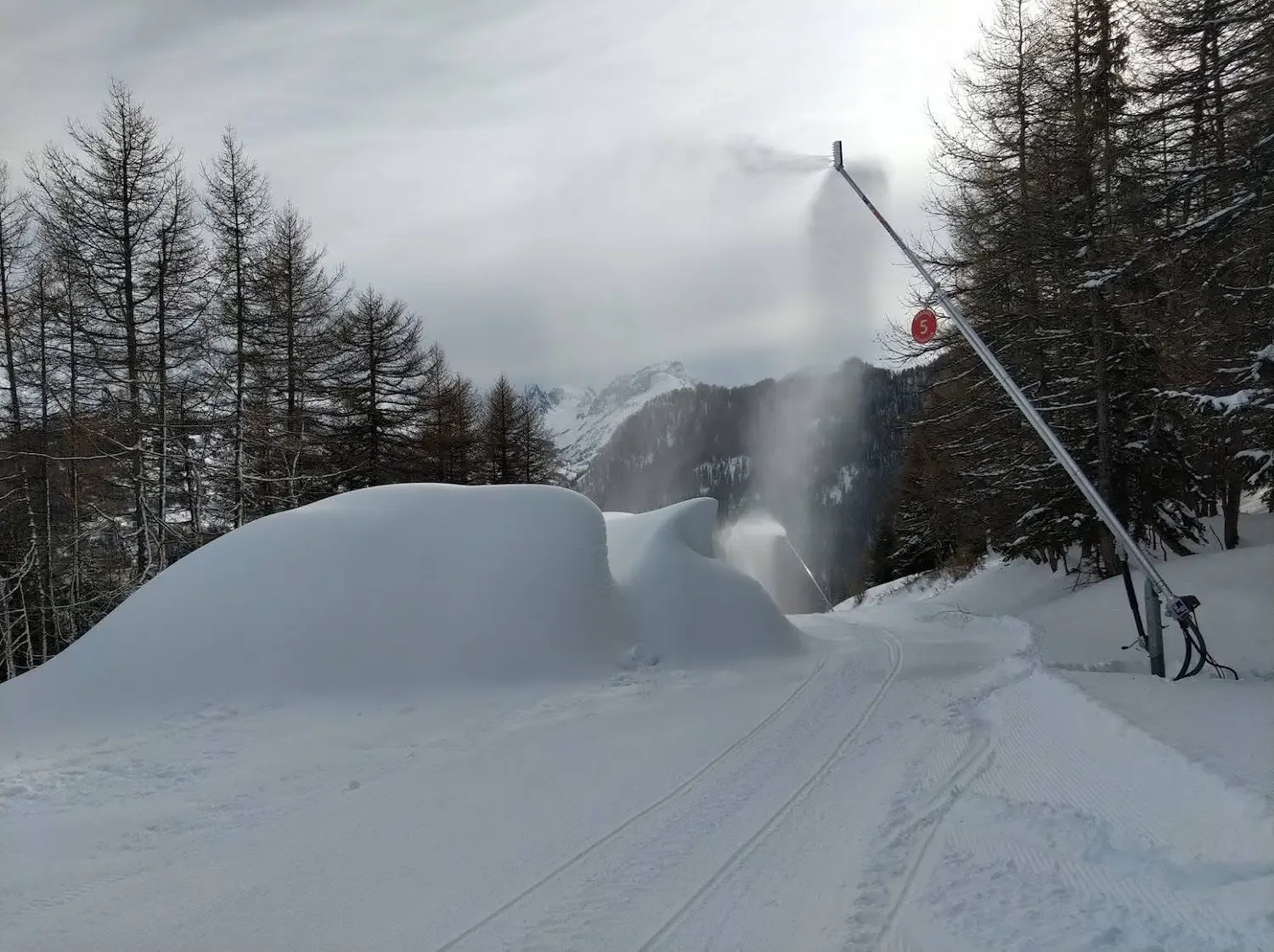 Snow-covered ski trail with a snowmaking machine covering the area in snow, surrounded by leafless trees and mountain peaks in the background.