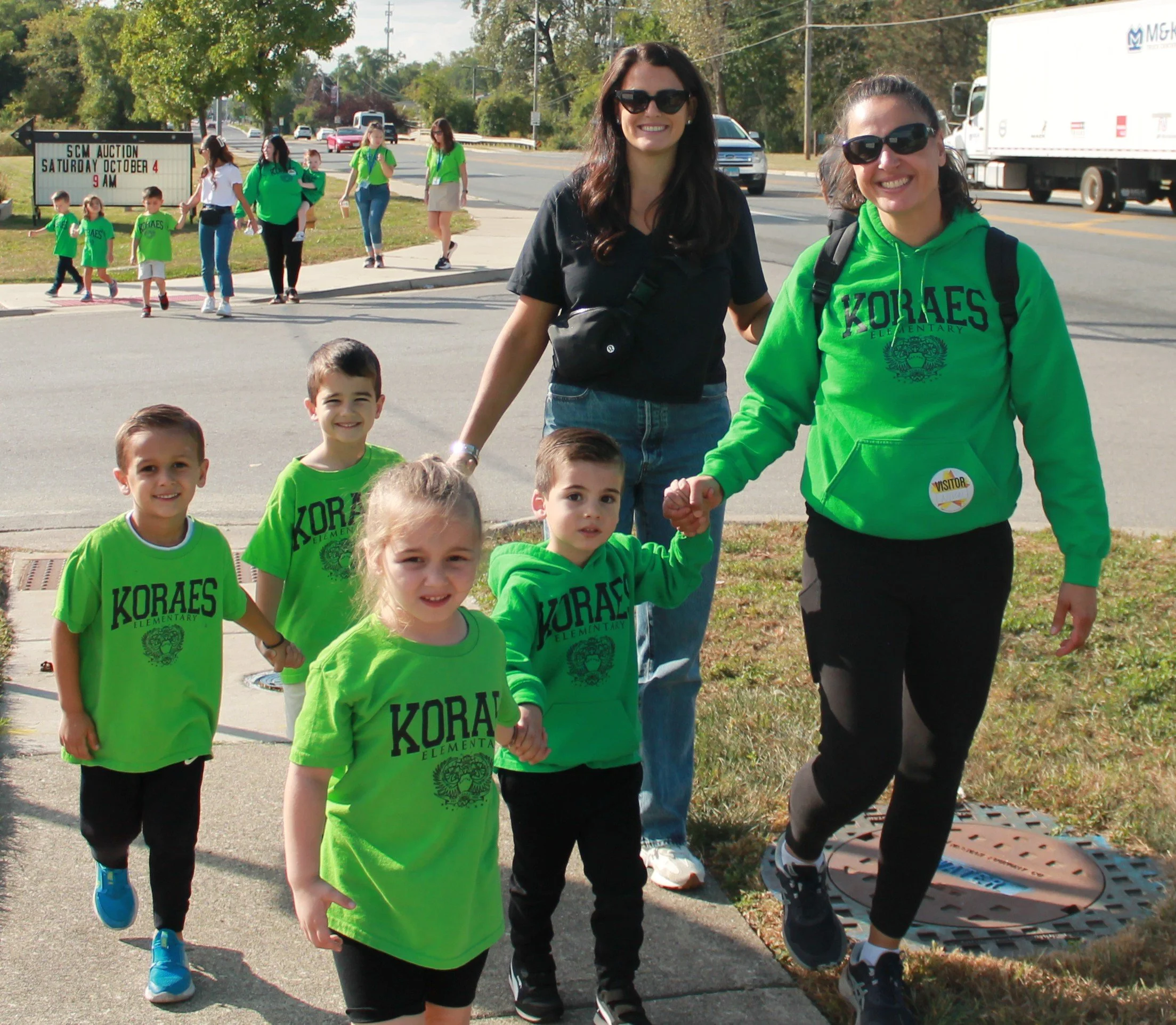 A group of children and adults walking outdoors, with children wearing bright green KORAES elementary shirts, and two women holding hands with children, smiling. In the background, more people and a road with moving trucks and cars.