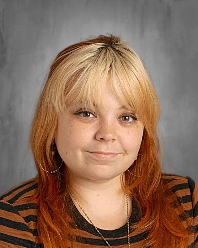 Portrait of a young woman with blonde hair and reddish-blonde highlights, wearing a striped brown and black top, against a plain gray background.