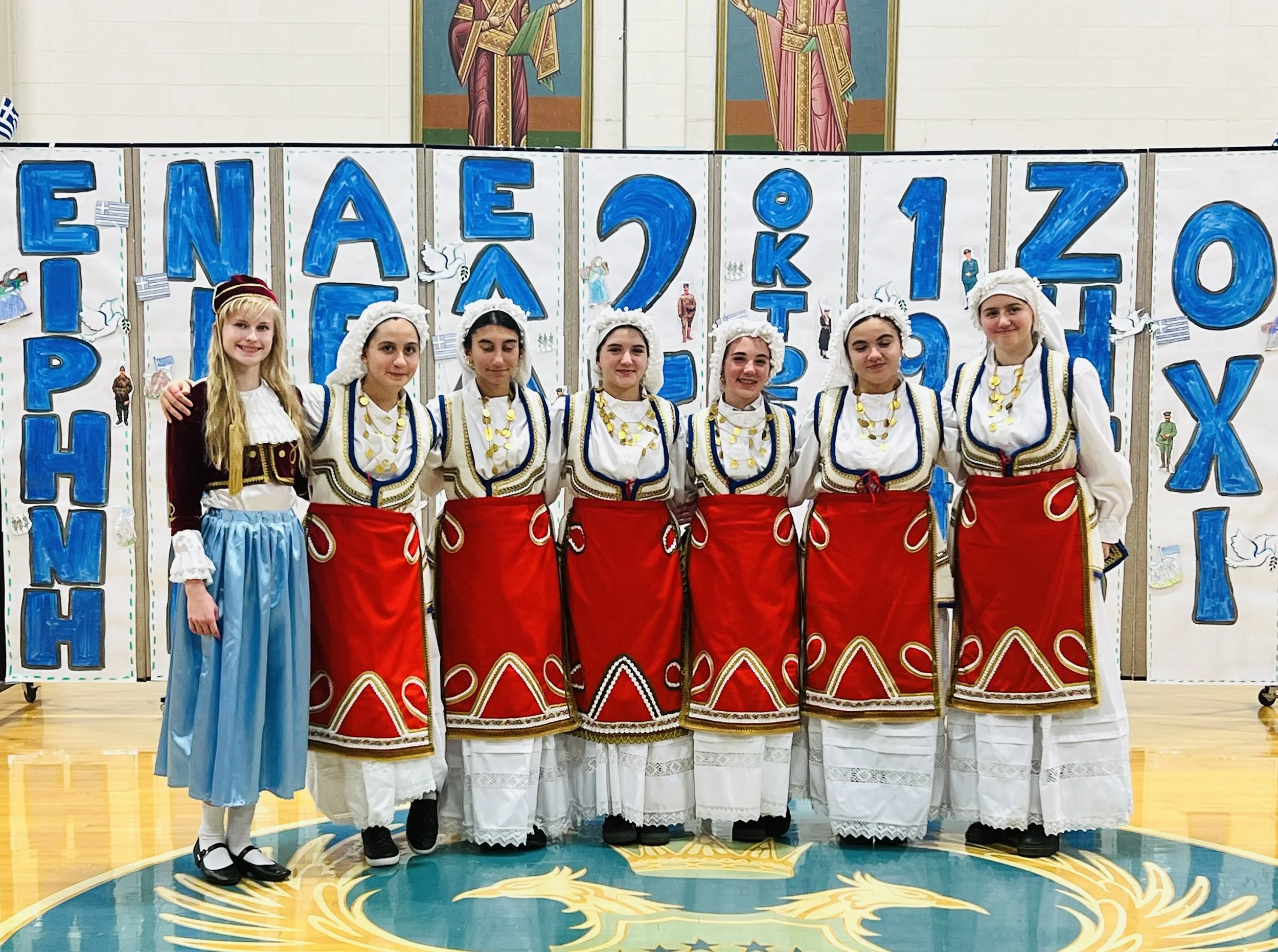 Group of young women in traditional Greek costumes standing in front of a festive backdrop with blue lettering and illustrations, inside a gymnasium.