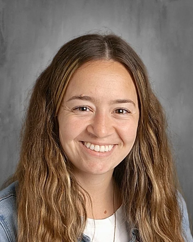 A young woman with long, wavy brown hair and a bright smile, wearing a white top, standing against a neutral gray background.