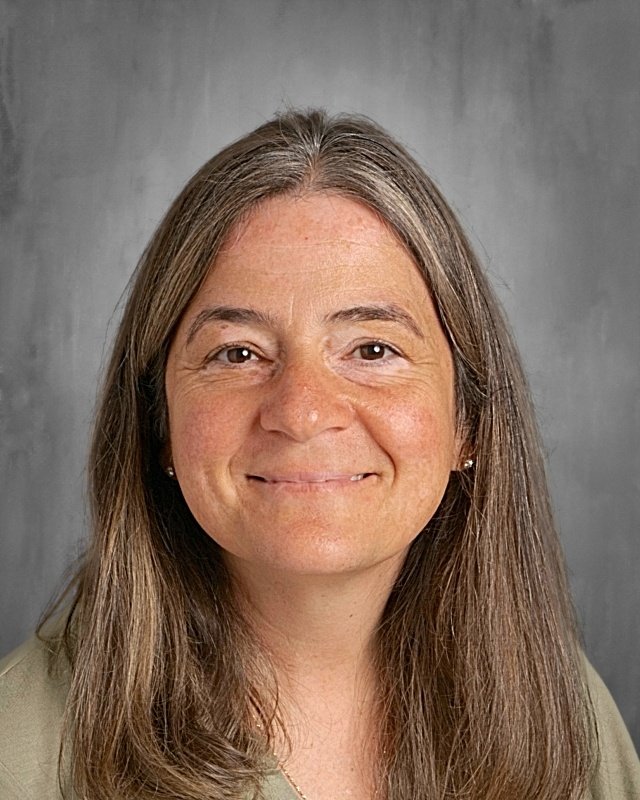 A woman with long, straight, brown hair smiling at the camera against a plain gray background.