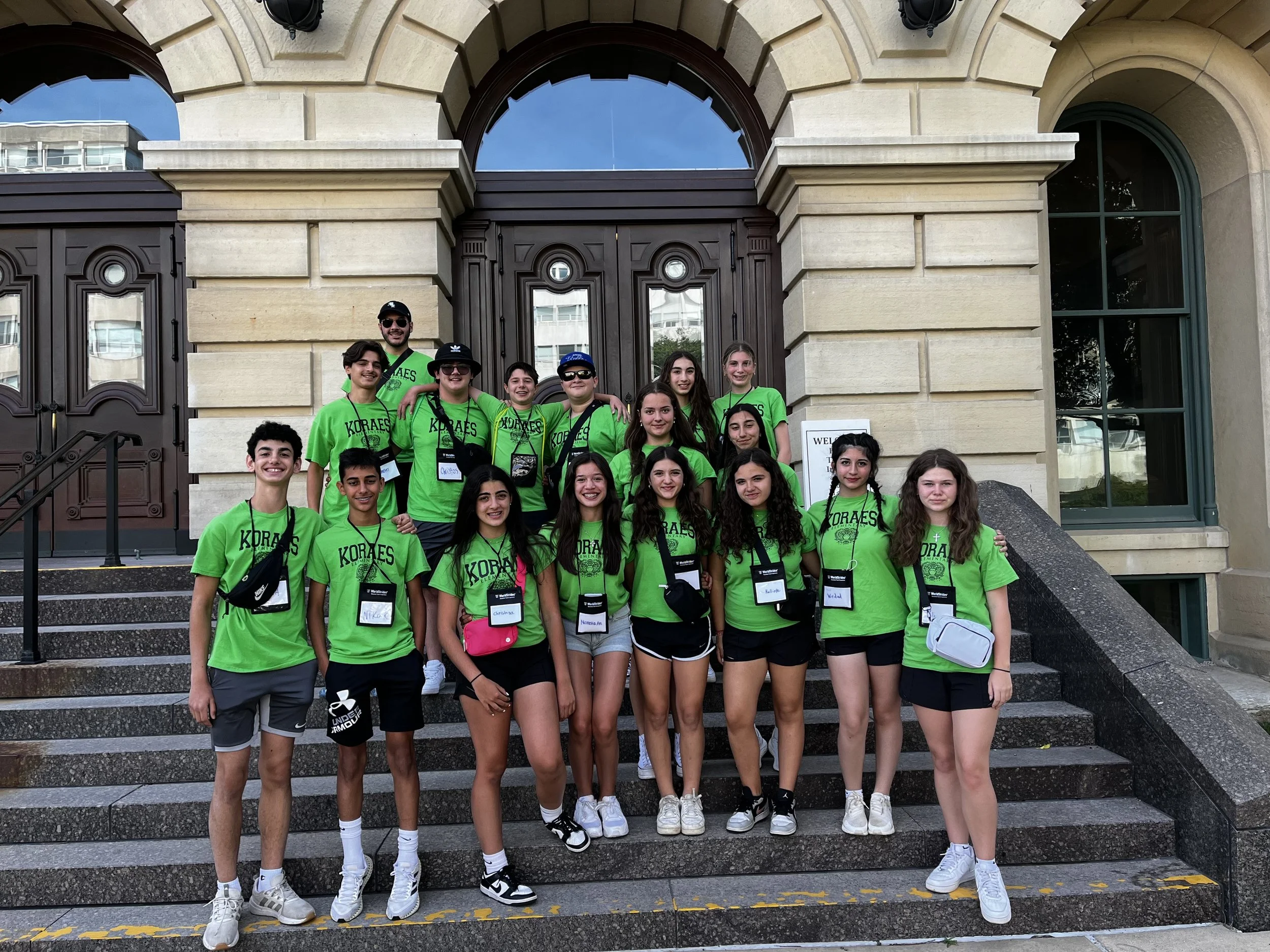 A group of young people wearing green t-shirts with 'KORAES' printed on them, standing on the steps of a historic building with large wooden doors and arched windows.