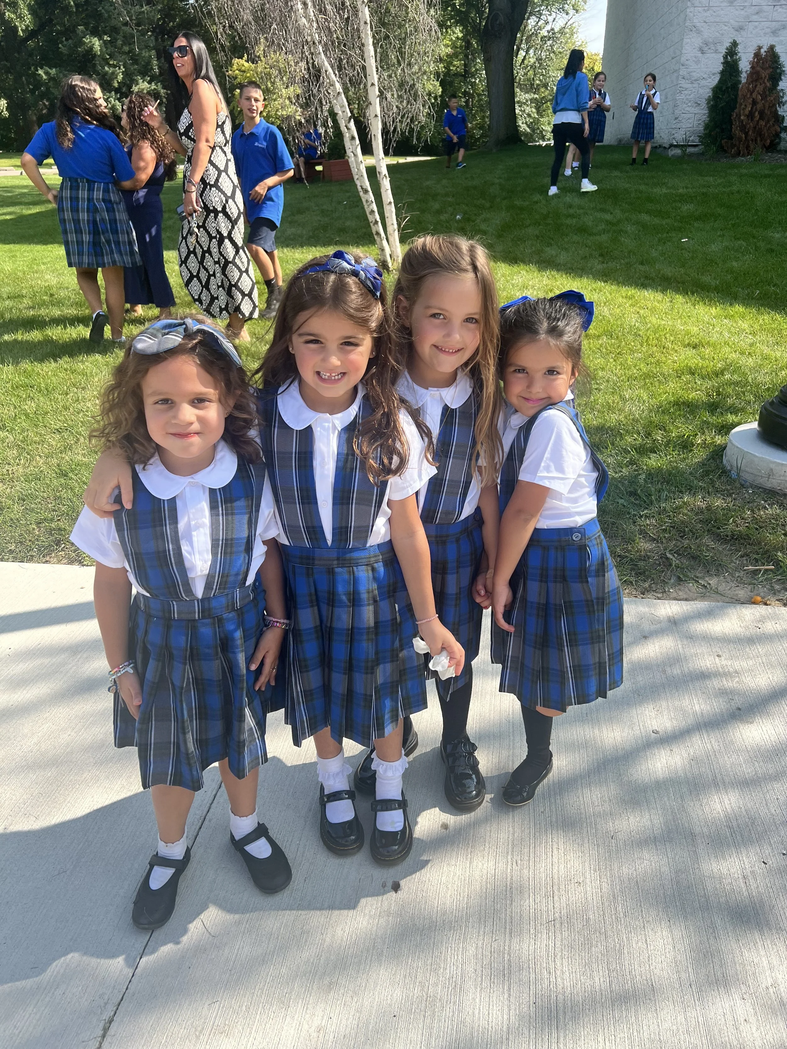 Four young girls in school uniforms smiling and posing together outside on a sidewalk with a grassy area, trees, and other children in the background.