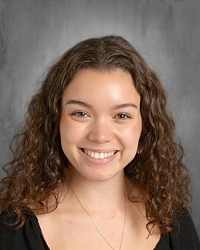 Portrait of a young woman with curly brown hair and a bright smile, wearing a black top and a delicate necklace.