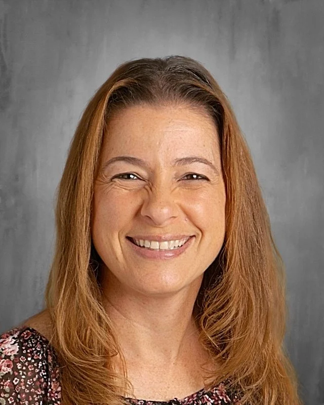 Portrait of a smiling woman with long, wavy, light brown hair, wearing a dark, patterned top against a gray background.