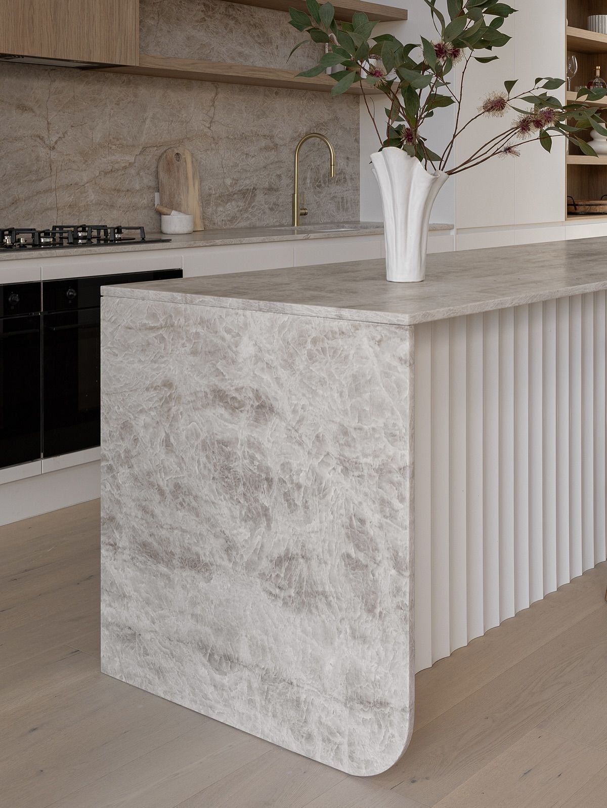 Modern kitchen with a beige marble island, a white vase with greenery, and a built-in stovetop with black cabinetry.