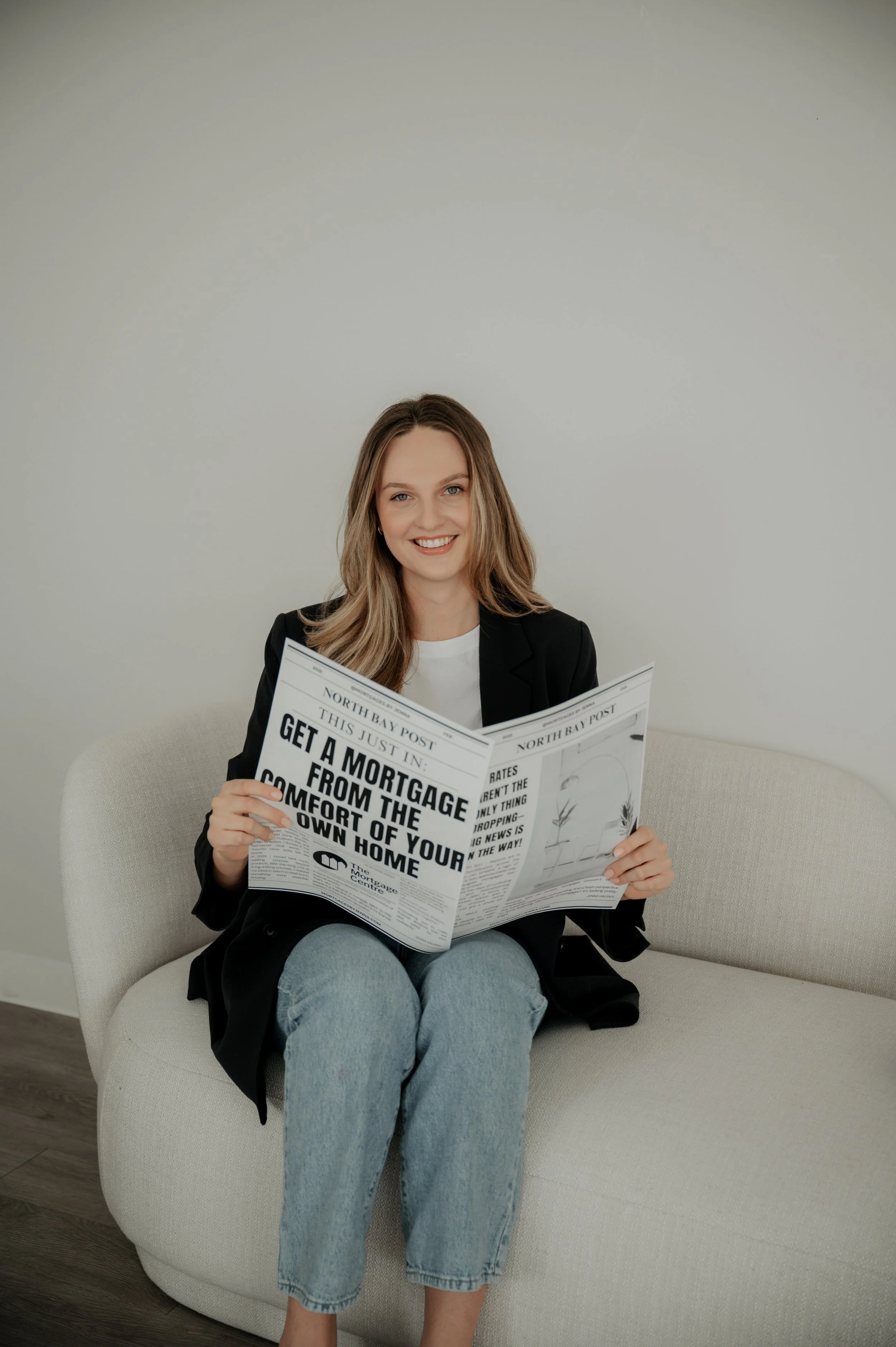 Jenna Valenti, mortgage agent,  wearing a black blazer and jeans, sitting on a light-colored couch, smiling, and reading a newspaper.
