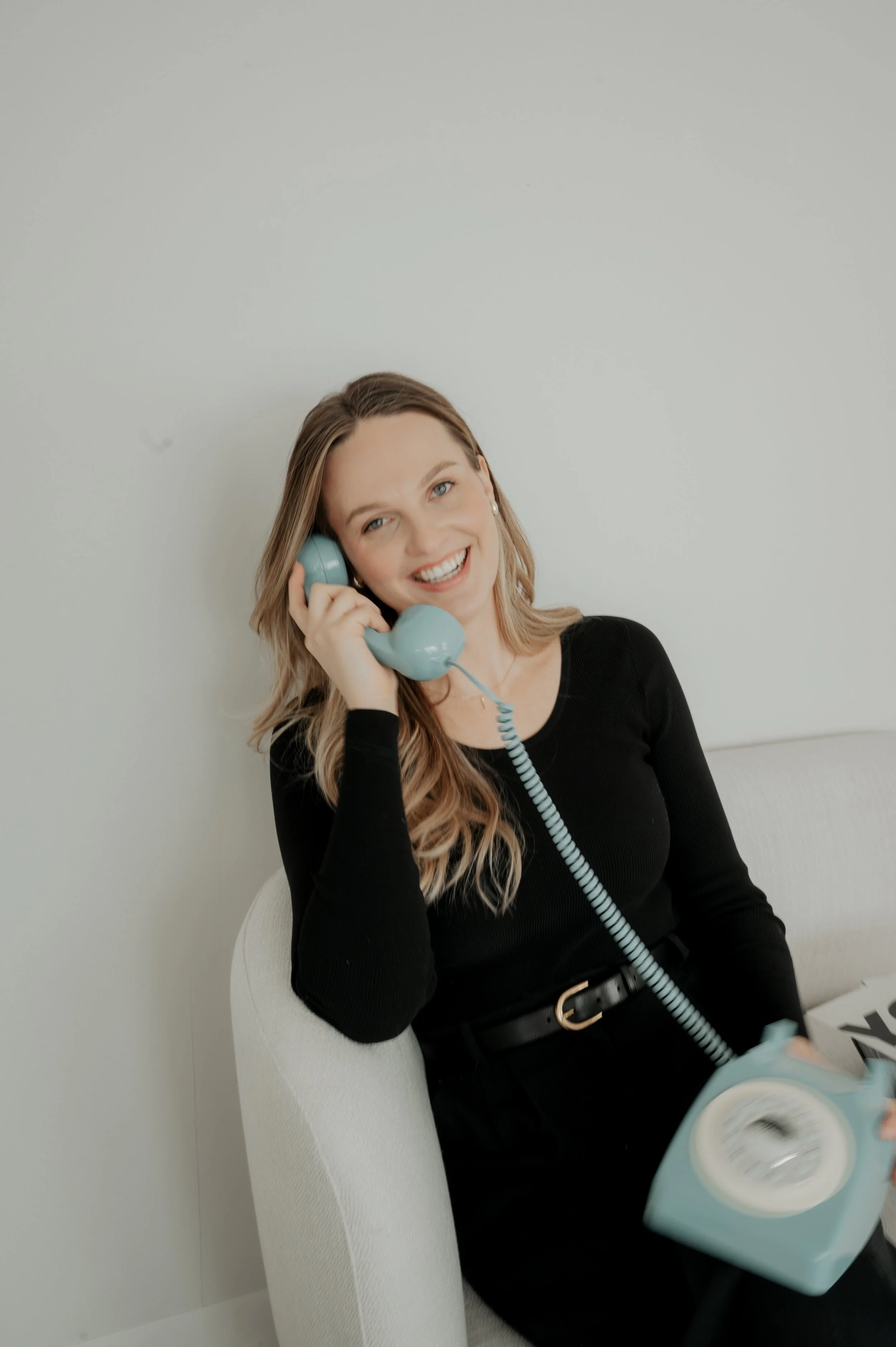 A smiling woman with blonde hair wearing a black top, sitting on a light-colored chair, while holding a vintage blue rotary phone receiver to her ear and a matching phone base in her lap.