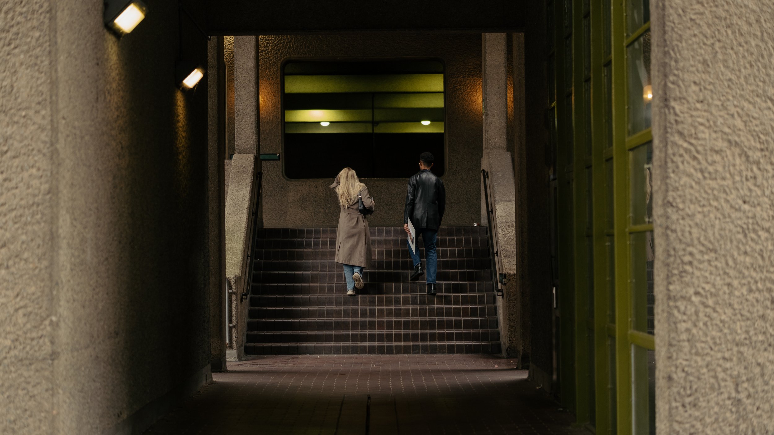 Two people ascending dark-colored stairs in an underpass, with textured beige walls, escorted by railings on each side, leading to a bright lobby or entrance area visible through an opening.