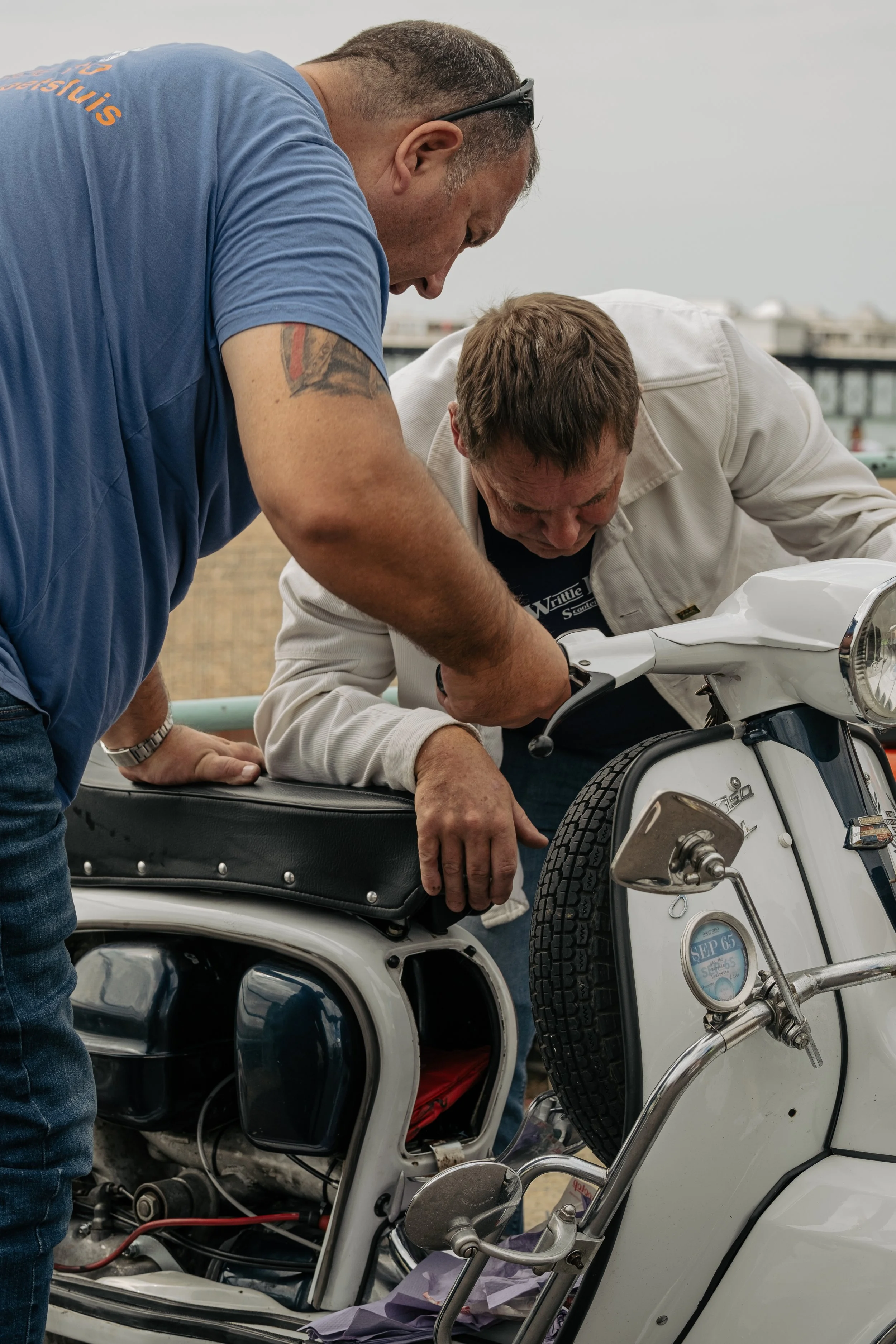 Two men working on a vintage white scooter, seemingly performing maintenance or repairs outdoors.