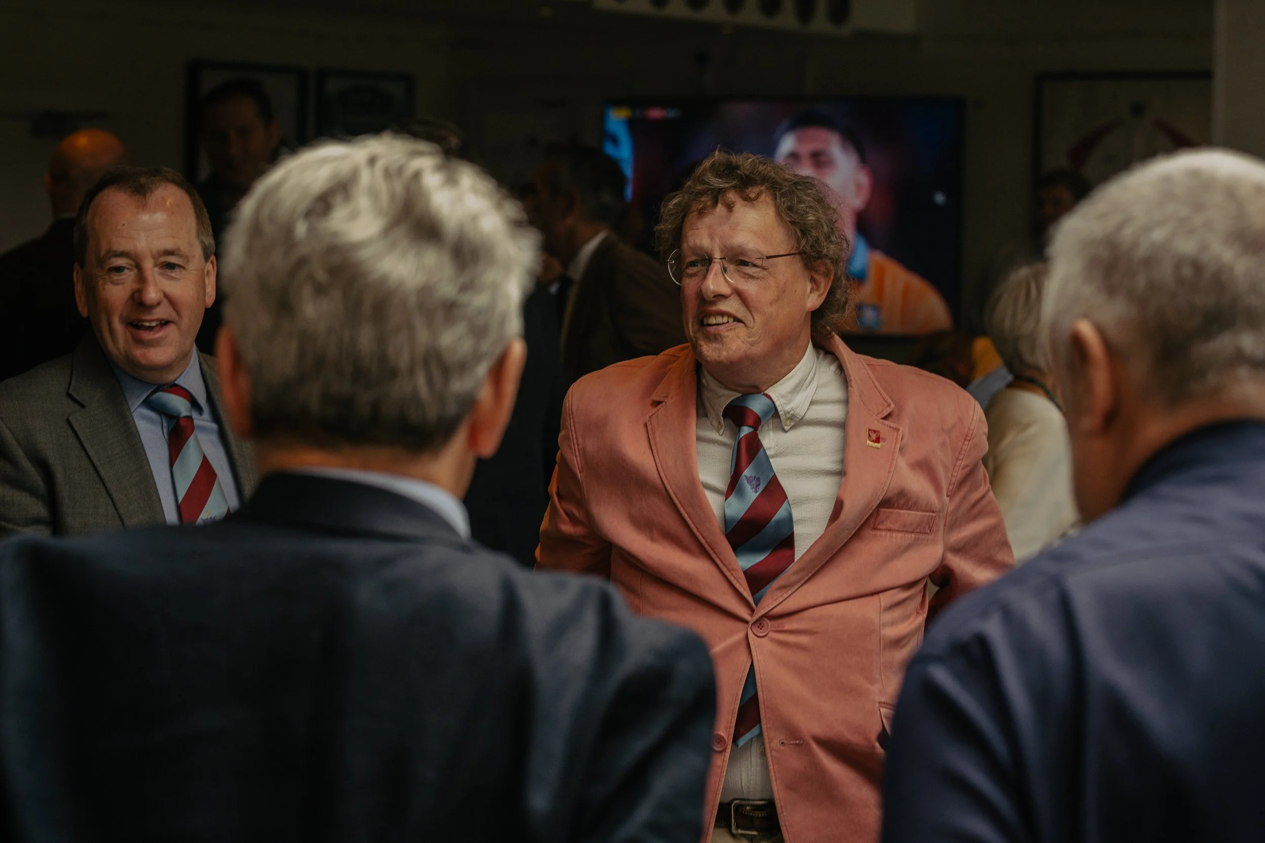 A group of men in business attire engaged in conversation at an event. One man is wearing a coral blazer, white shirt, and a striped tie, and appears to be speaking or reacting to something. The others are dressed in suits and are listening or talkin