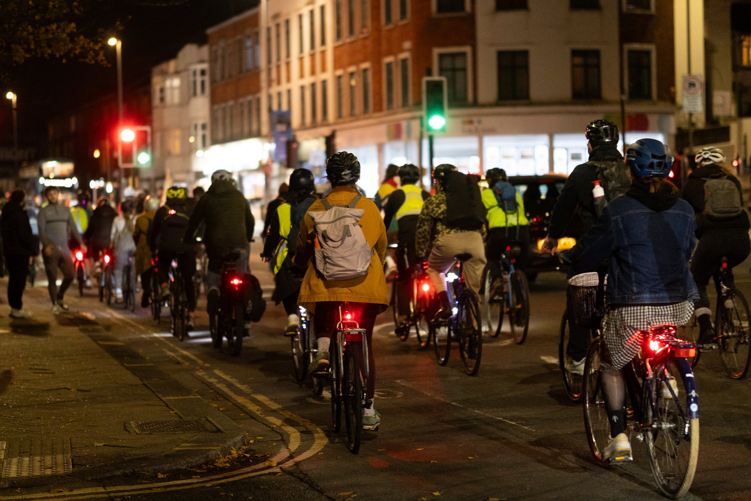 Group of people riding bicycles at night on a busy city street, some wearing helmets, illuminated by streetlights and bicycle lights, with buildings and traffic signals in the background.