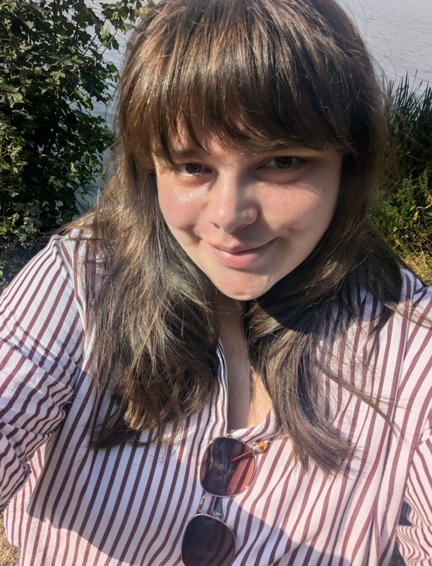 A woman with shoulder-length brown hair and bangs smiling outdoors near a body of water, wearing a striped button-up shirt with sunglasses hanging from the collar.
